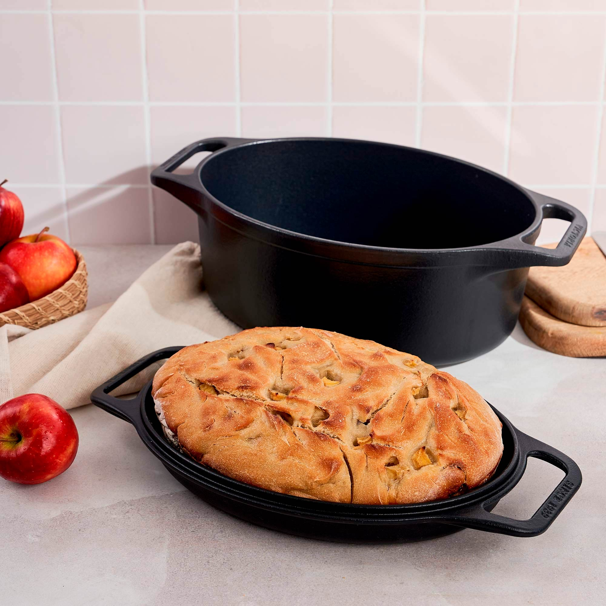 Pan en un plato negro, manzanas y una olla negra sobre una mesa. Pared de baldosas rosas al fondo.