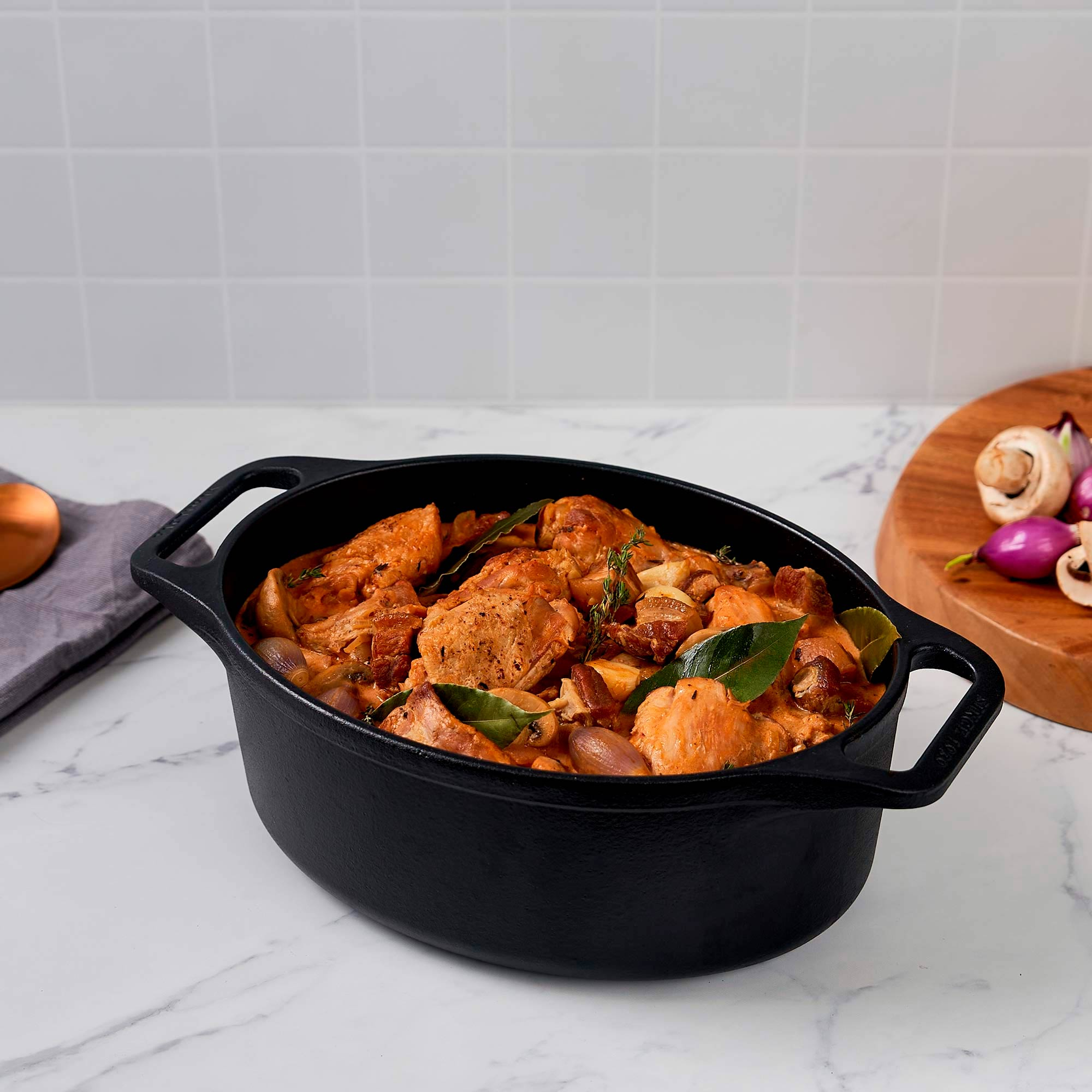Una olla de hierro fundido con estofado de pollo, chalotes y champiñones. La olla está sobre una superficie de mármol, junto a una tabla de madera.