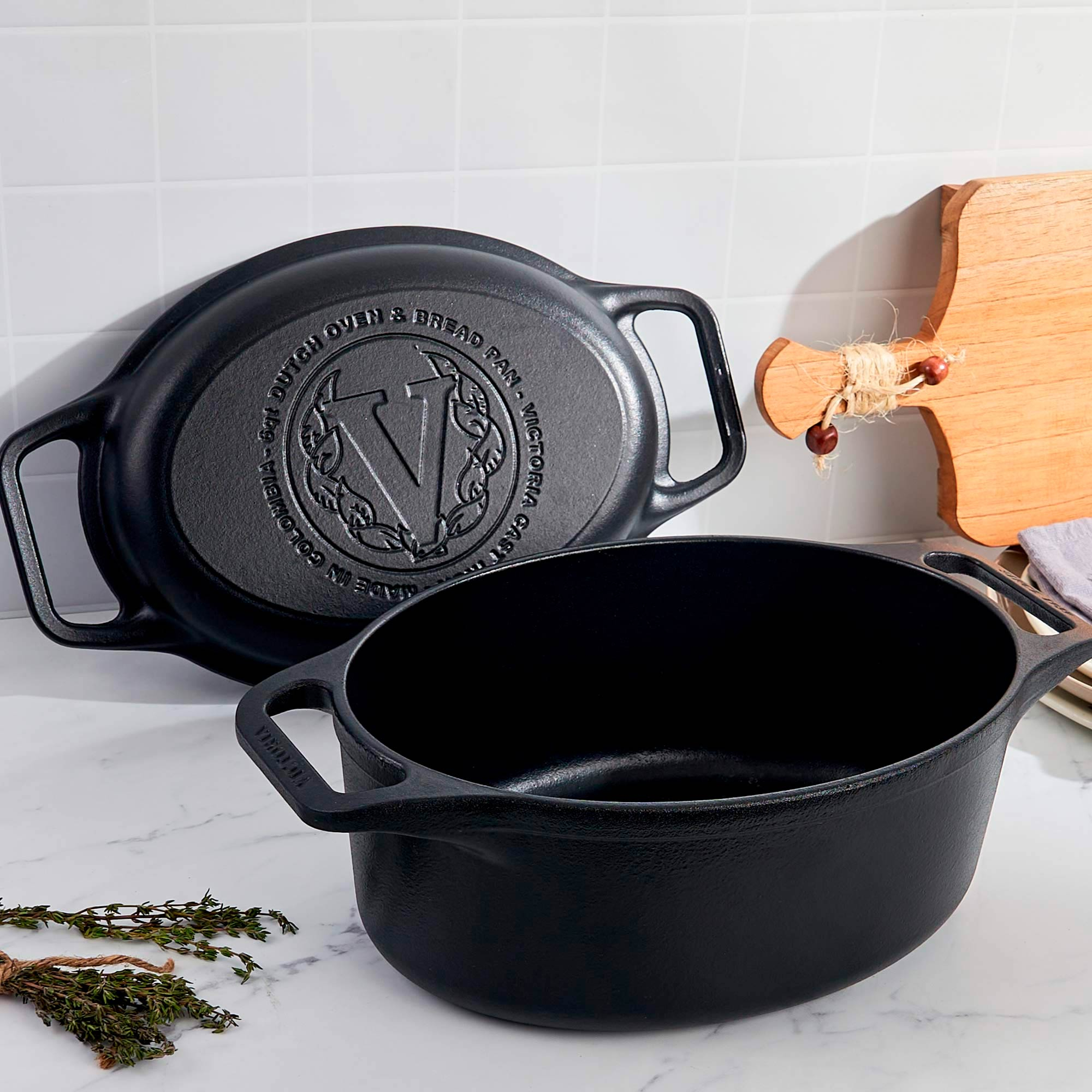 Olla holandesa de hierro fundido negro con tapa. Tabla de cortar de madera al fondo. Hierbas frescas a un lado.