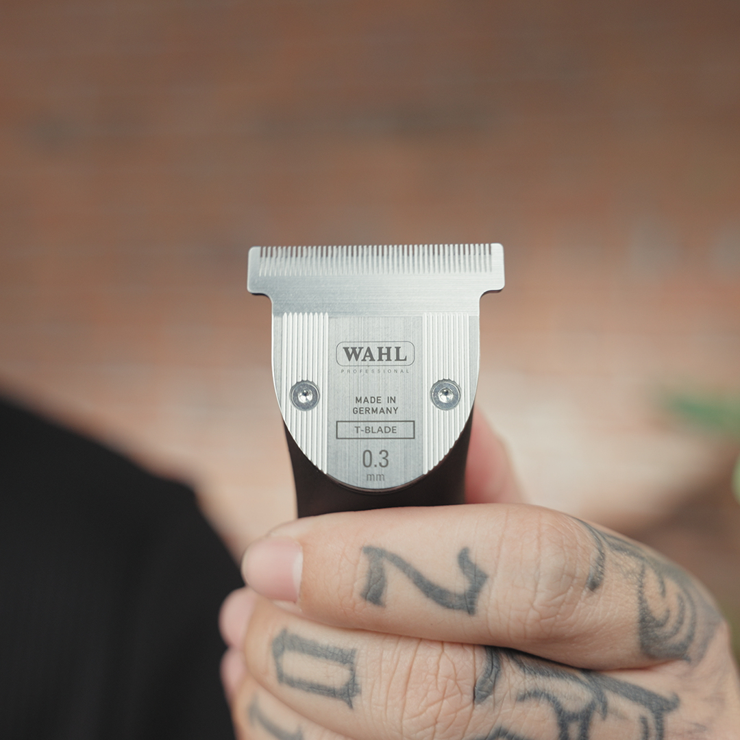 Une main tient une tondeuse Wahl T-blade. La lame est argentée avec le logo et le texte Wahl. La main est tatouée et sur un fond flou.