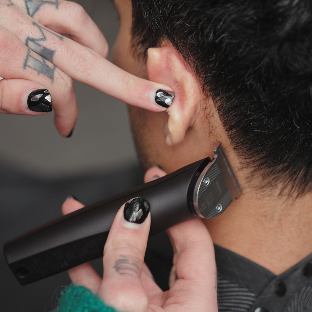 Les cheveux d'une personne sont coupés avec une tondeuse. La main de la personne avec du vernis à ongles noir tient la tondeuse.