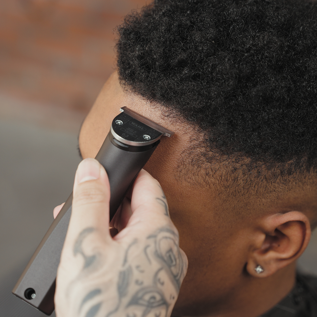 Un coiffeur utilise une tondeuse sur la ligne de cheveux d'un homme noir. La main tatouée du coiffeur tient la tondeuse. L'homme porte une boucle d'oreille.