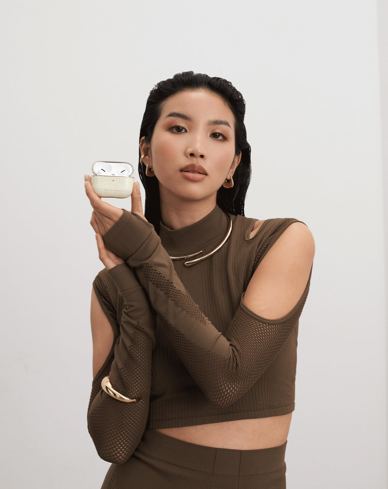 Eine Frau hält ein offenes AirPods-Etui, trägt ein braunes Outfit mit Goldschmuck, vor weißem Hintergrund.