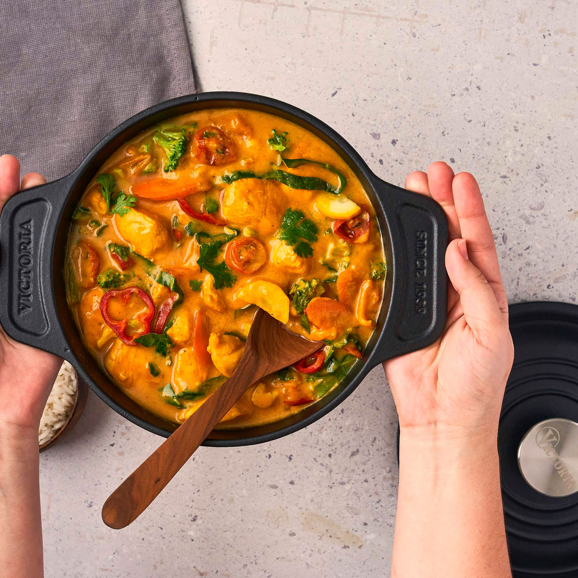 Vista cenital de una olla negra con curry, sostenida por manos. Se ven verduras, una cuchara de madera y arroz.