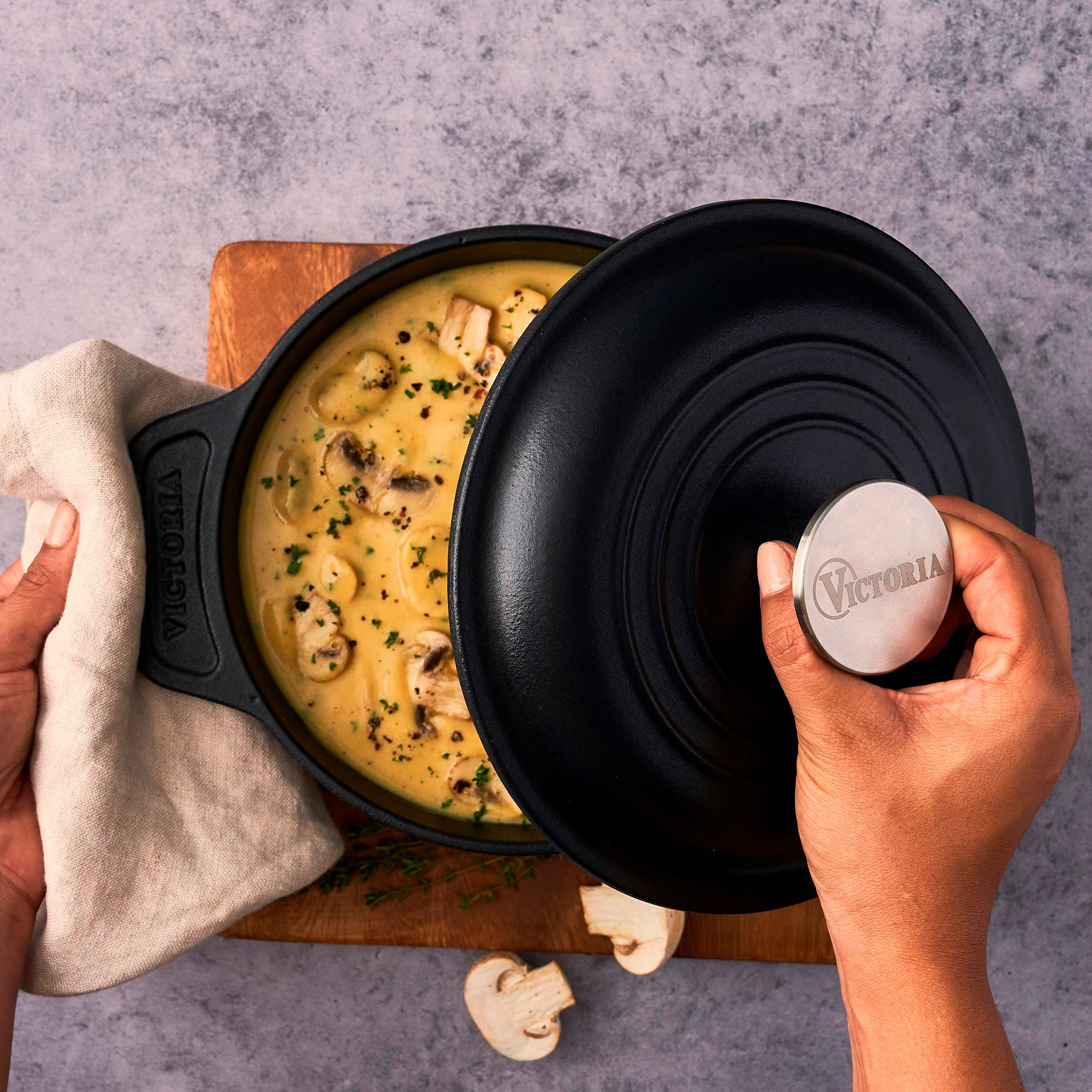 Una persona abre una olla negra de sopa de champiñones. La olla está sobre una tabla de madera. Manos sosteniendo un paño y la tapa.