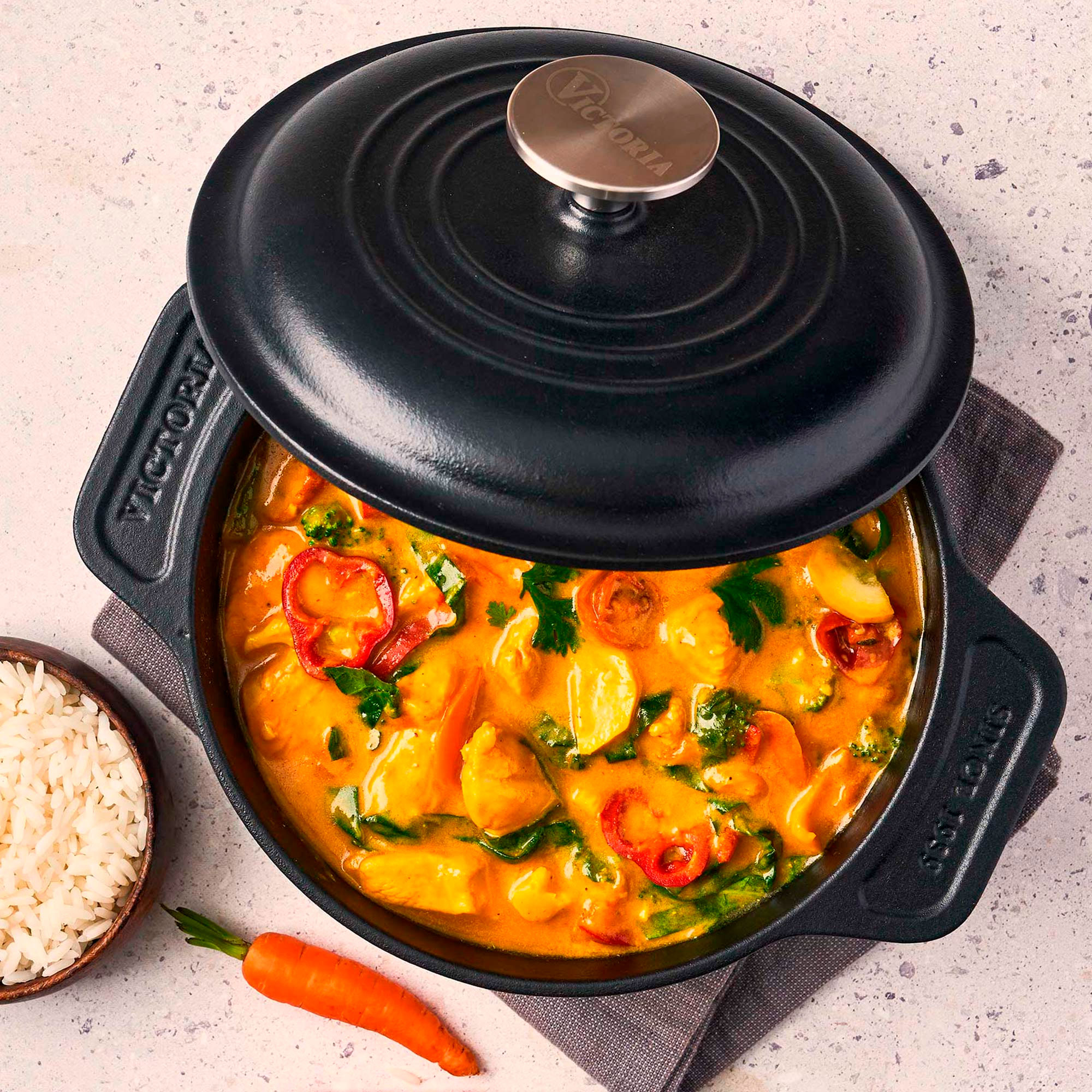 Olla negra con curry y verduras, un cuenco de arroz y una zanahoria sobre piedra.