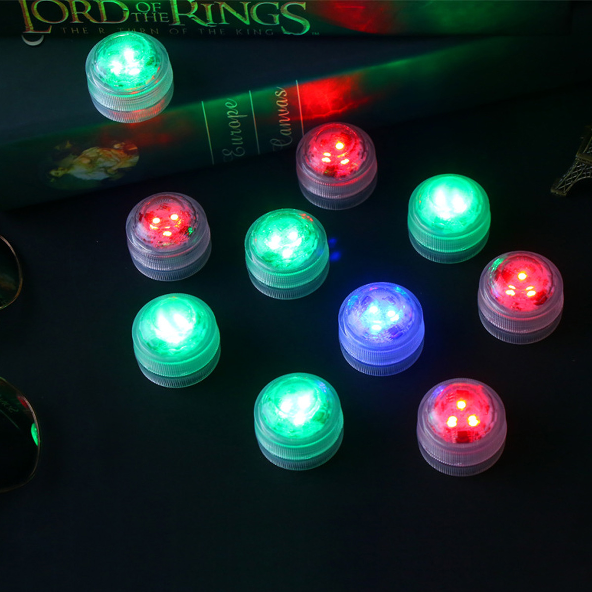 Mehrere kleine, beleuchtete Lichter in verschiedenen Farben (Grün, Rot, Blau) sind auf einer dunklen Oberfläche angeordnet, in der Nähe eines Buches.