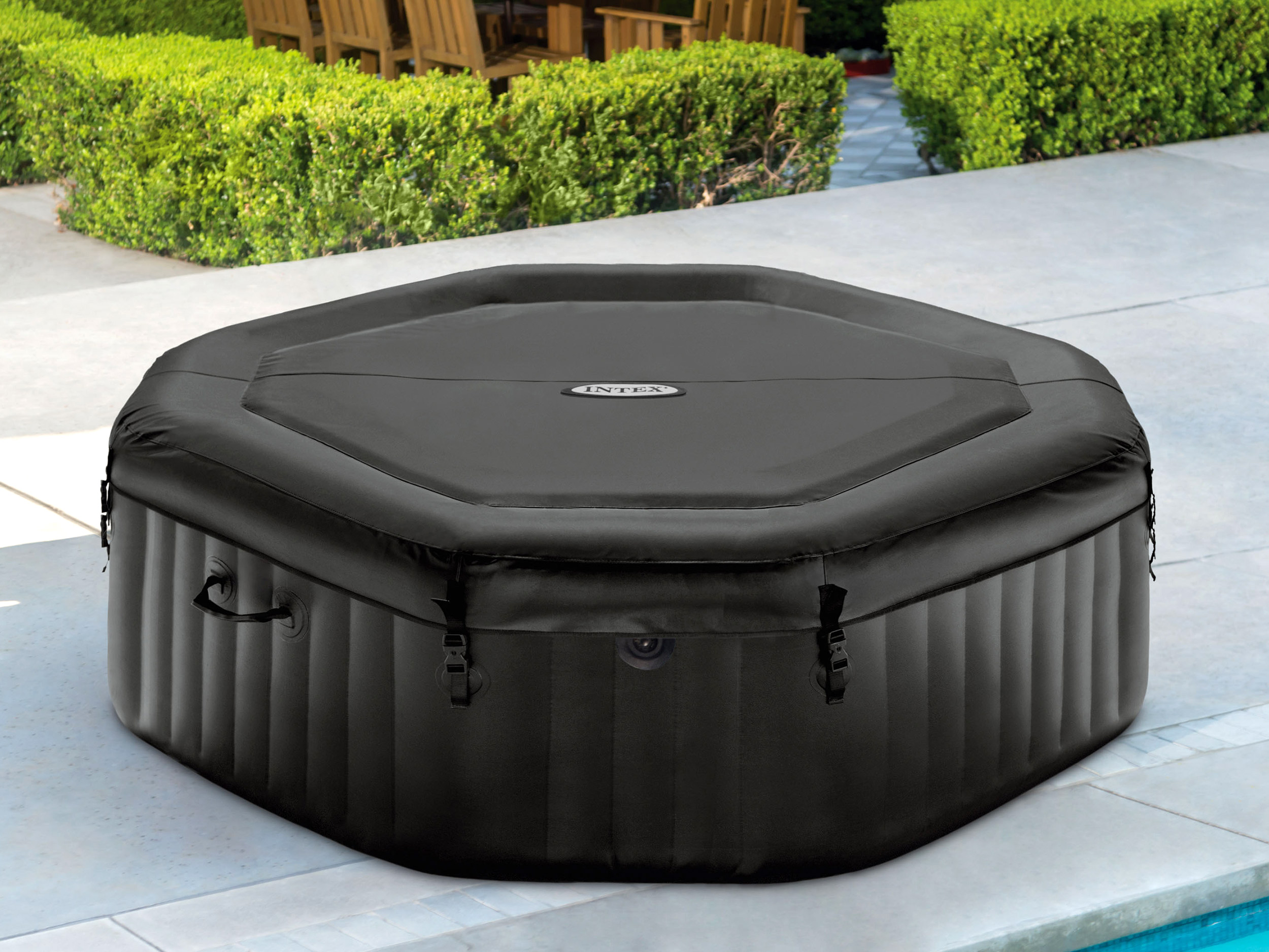 Czarna, sześciokątna wanna z pokrywą, na patio. Zielone krzewy i drewniane krzesła w tle.