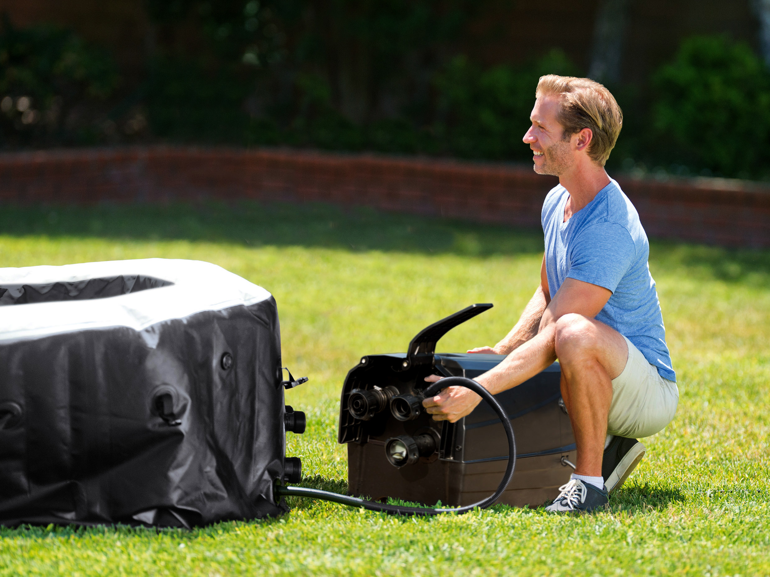 Mężczyzna w pobliżu nadmuchiwanej wanny z hydromasażem. Podłącza pompę. Zielona trawa i drzewa w tle.