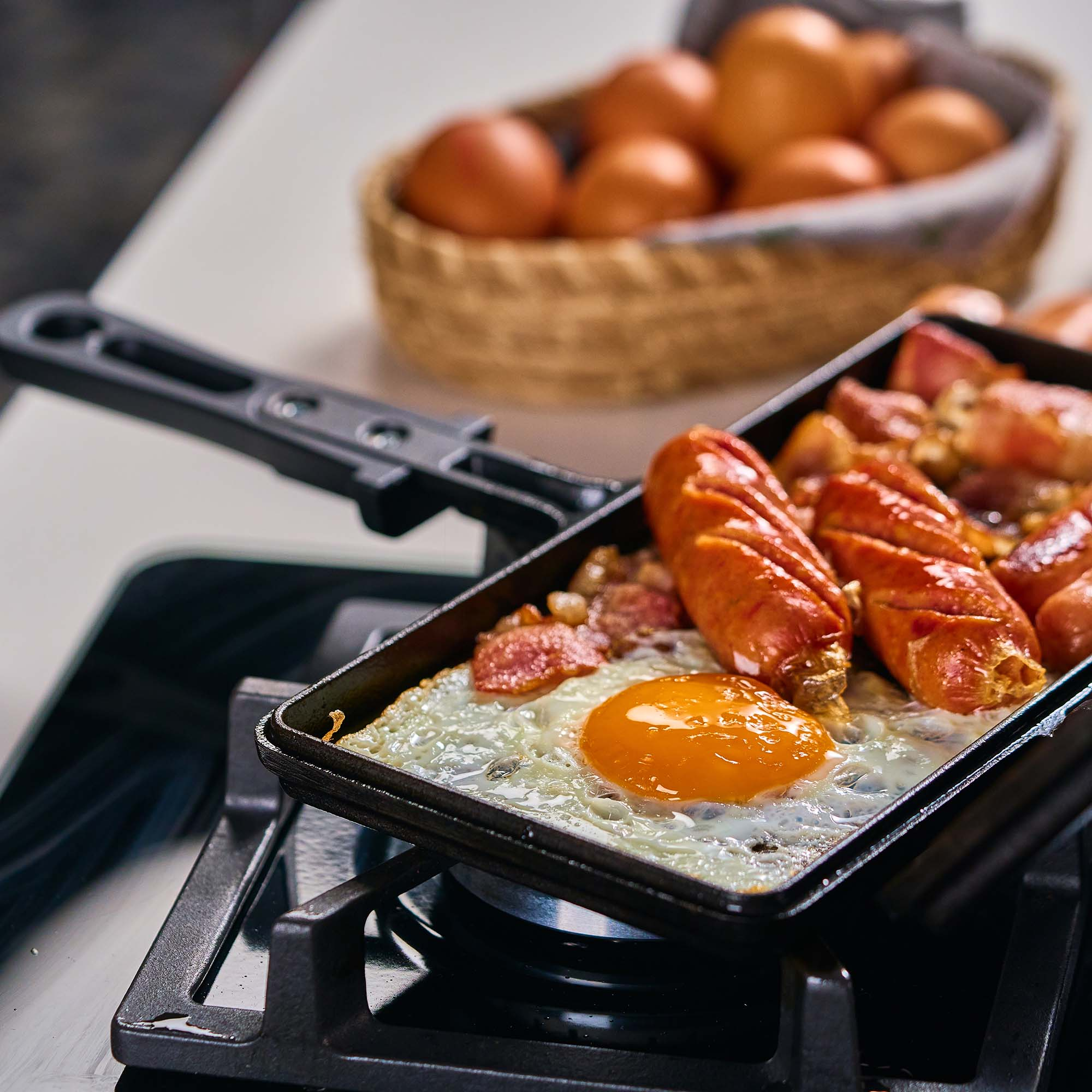 Una sartén con huevos fritos y salchichas en una estufa, con una cesta de huevos al fondo.