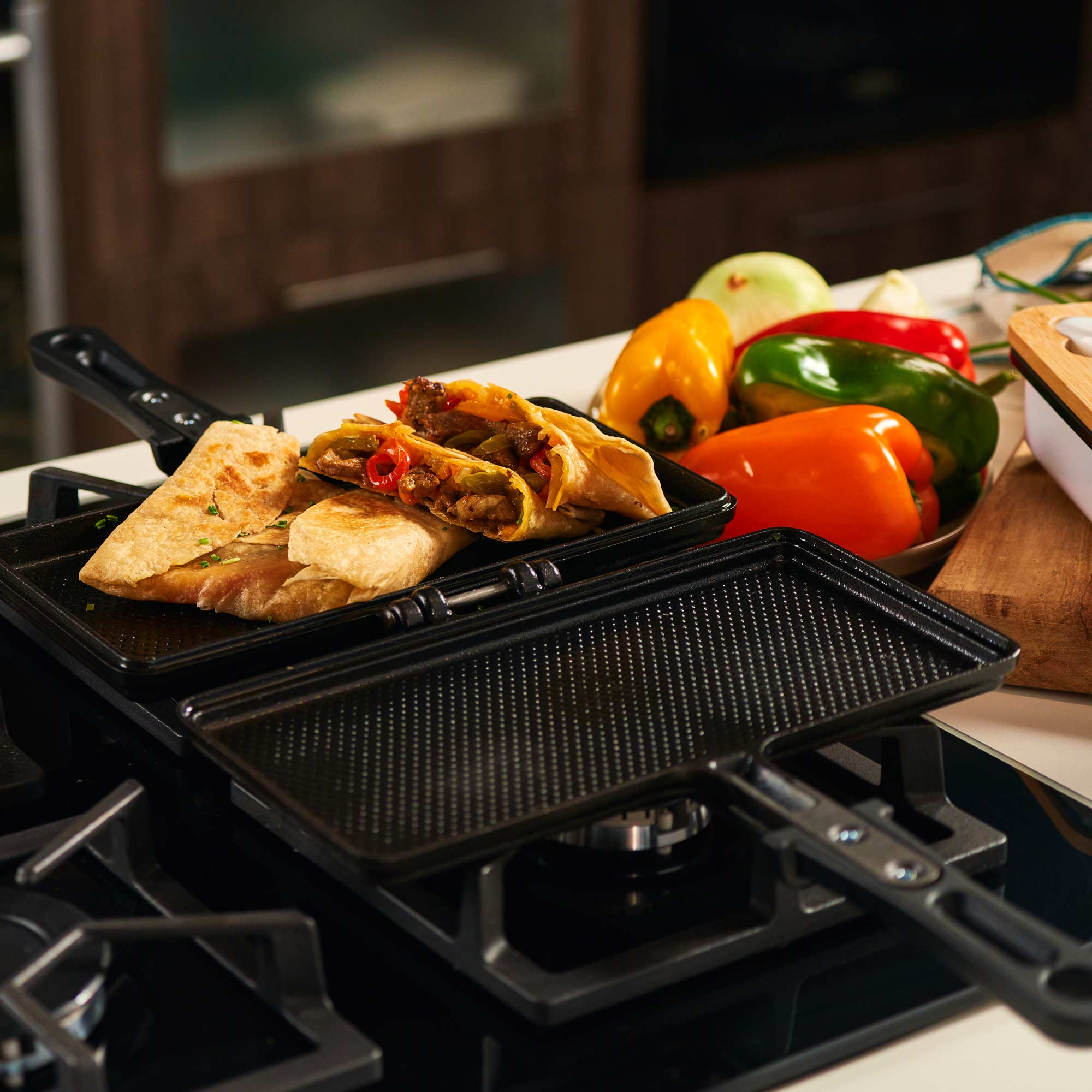 Una plancha para sándwiches con wraps rellenos. Pimientos y cebollas en el fondo. La plancha está sobre una estufa.