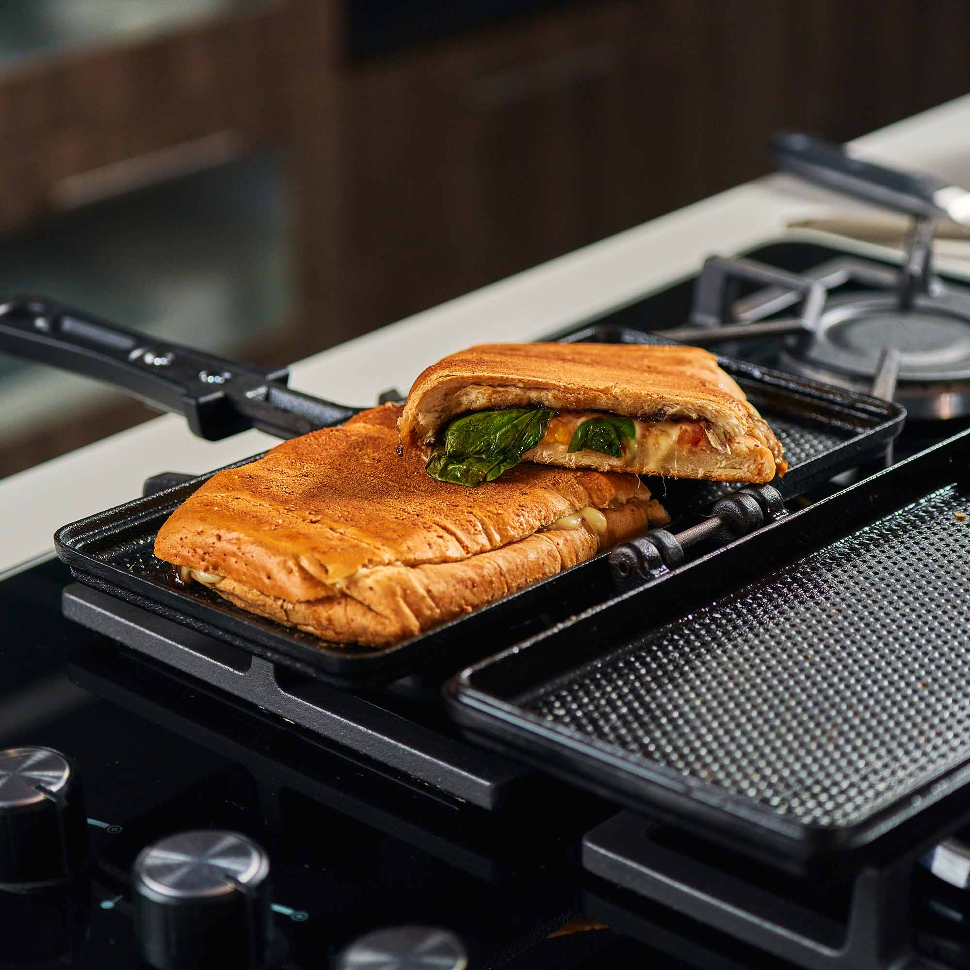 Un sándwich tostado cortado por la mitad, relleno de queso y espinacas, en una sandwichera negra sobre una estufa.