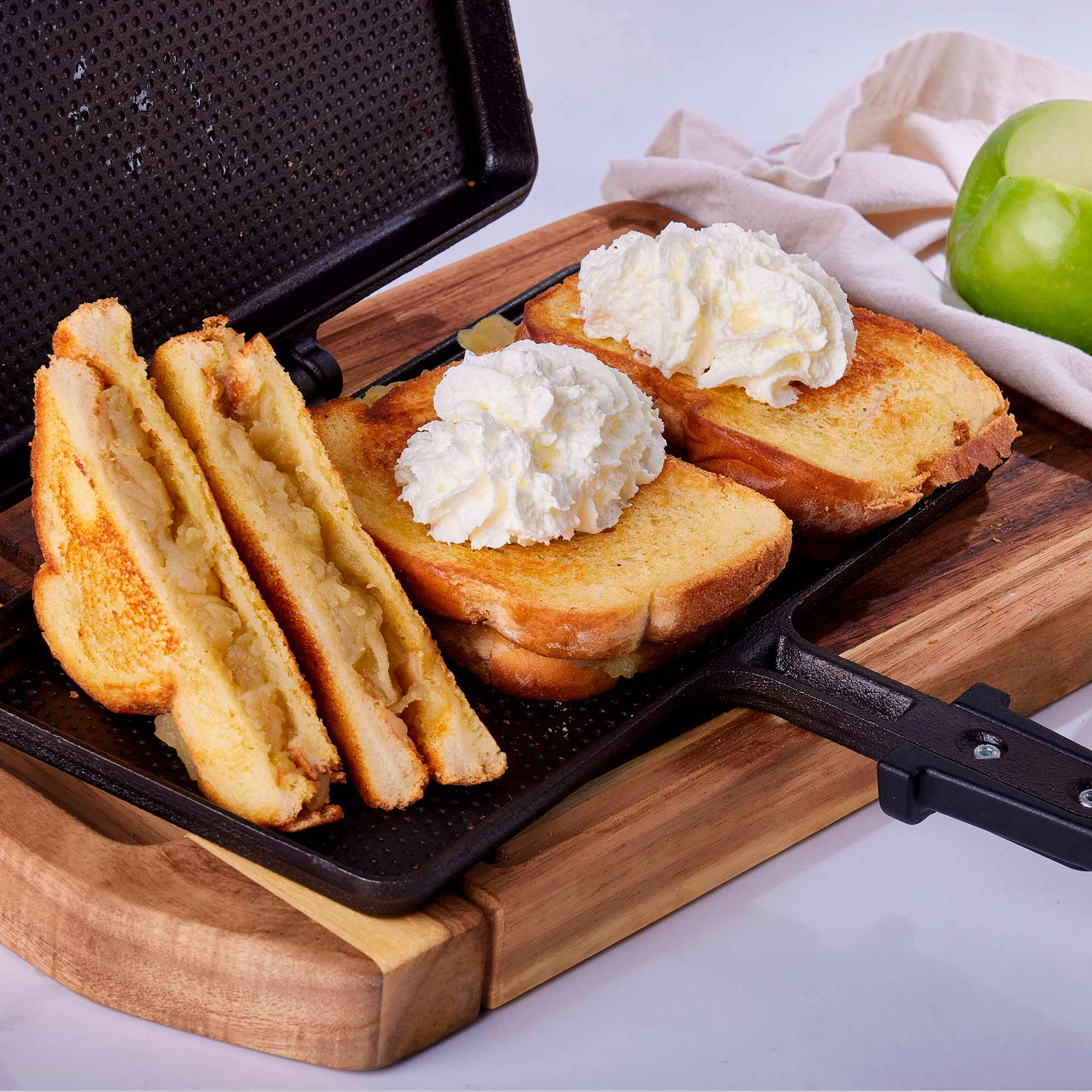 Sándwiches de manzana a la plancha con crema batida en una tabla de madera con una manzana verde y un paño.