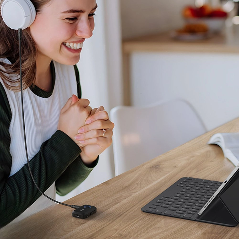 Uśmiechnięta kobieta w słuchawkach, korzystająca z tabletu z klawiaturą na drewnianym stole. Czarny gadżet podłączony do słuchawek.