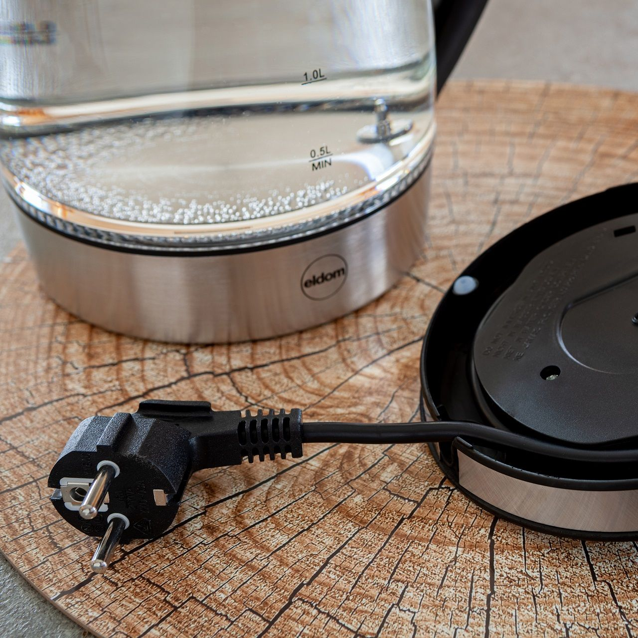 Un bollitore elettrico in vetro con spina nera su una superficie effetto legno. Il coperchio è aperto. Il bollitore ha acqua all'interno.