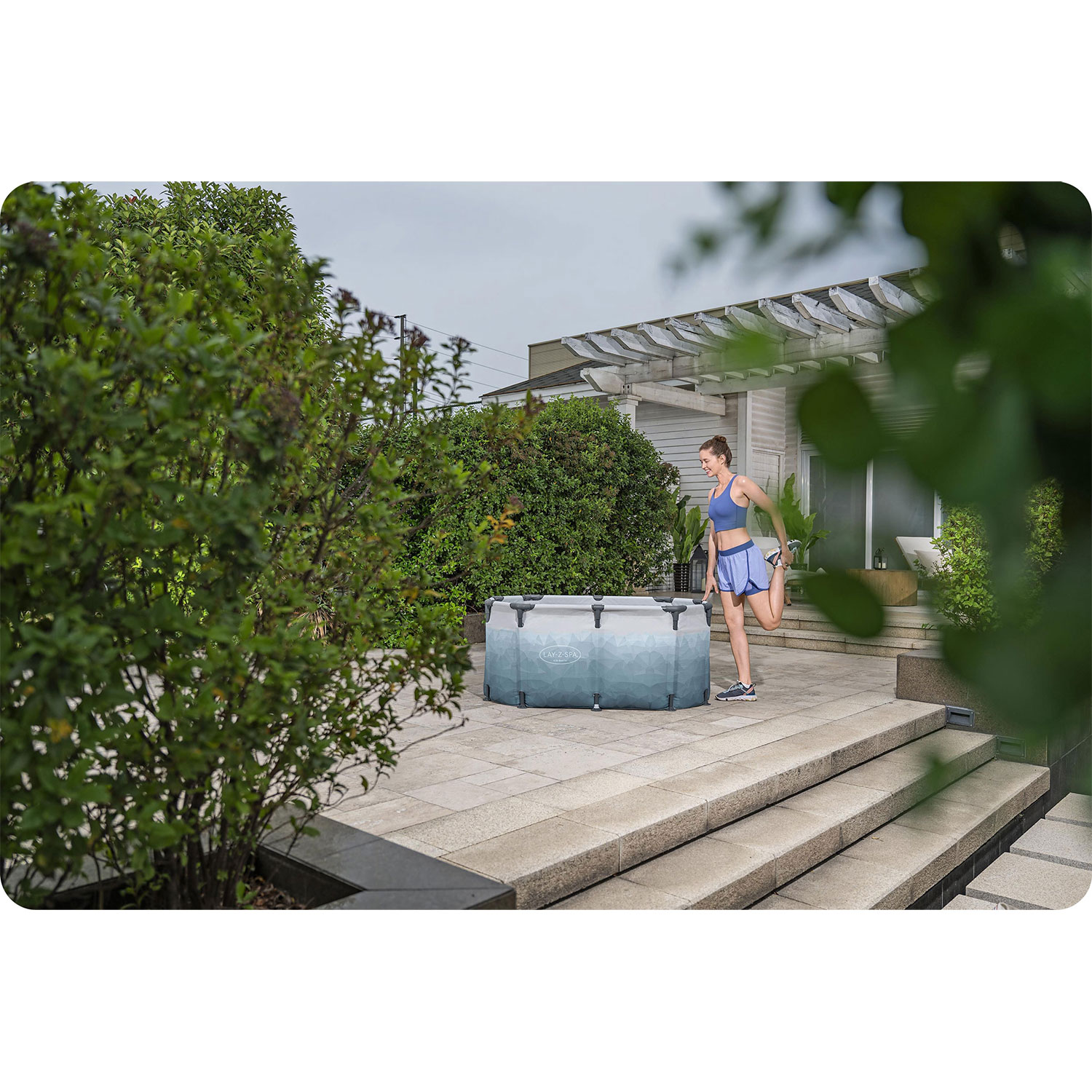 Kobieta rozciąga się w pobliżu wanny z hydromasażem. Ma na sobie niebieski stanik sportowy i szorty. Zielone krzewy i dom z pergolą w tle.