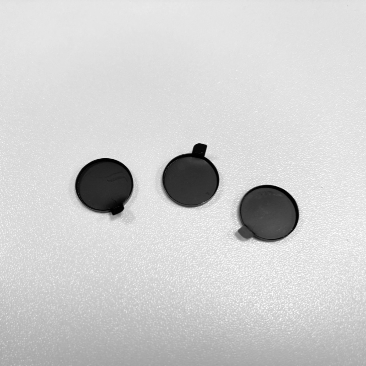 Trzy czarne, okrągłe obiekty z małymi zakładkami na białej, teksturowanej powierzchni. Są równomiernie rozmieszczone.