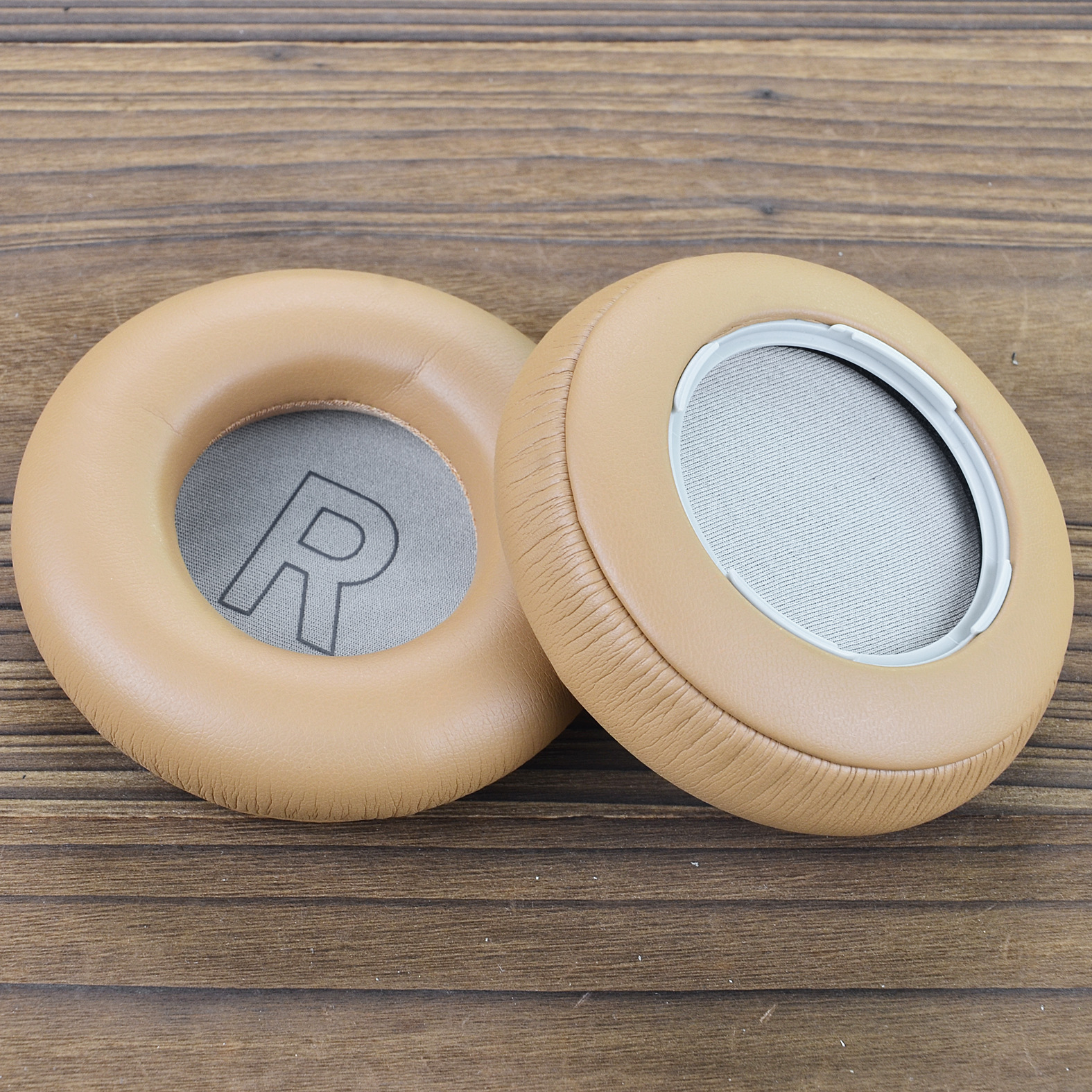 Dwie brązowe poduszki do słuchawek na drewnianej powierzchni. Jedna ma duże 'R'. Tło to drewno.