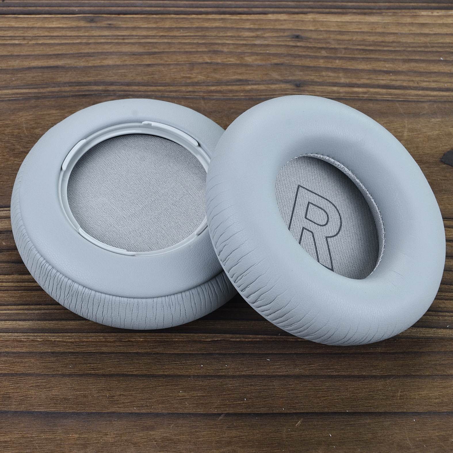 Dwie szare poduszki nauszne do słuchawek na drewnianej powierzchni. Jedna poduszka wyświetla literę 'R'.