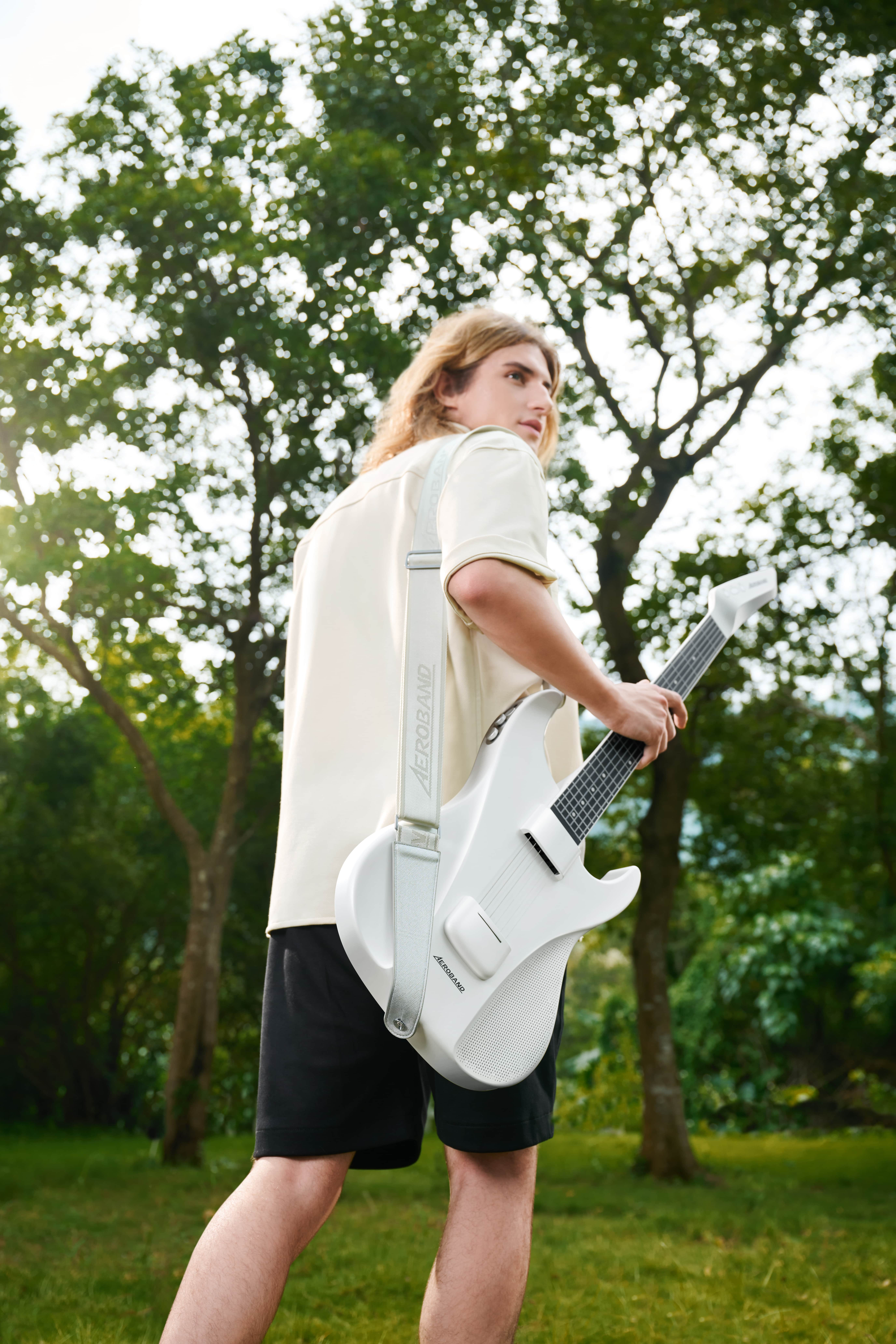 Een man met lang haar houdt een witte gitaar vast. Hij staat buiten met bomen op de achtergrond.