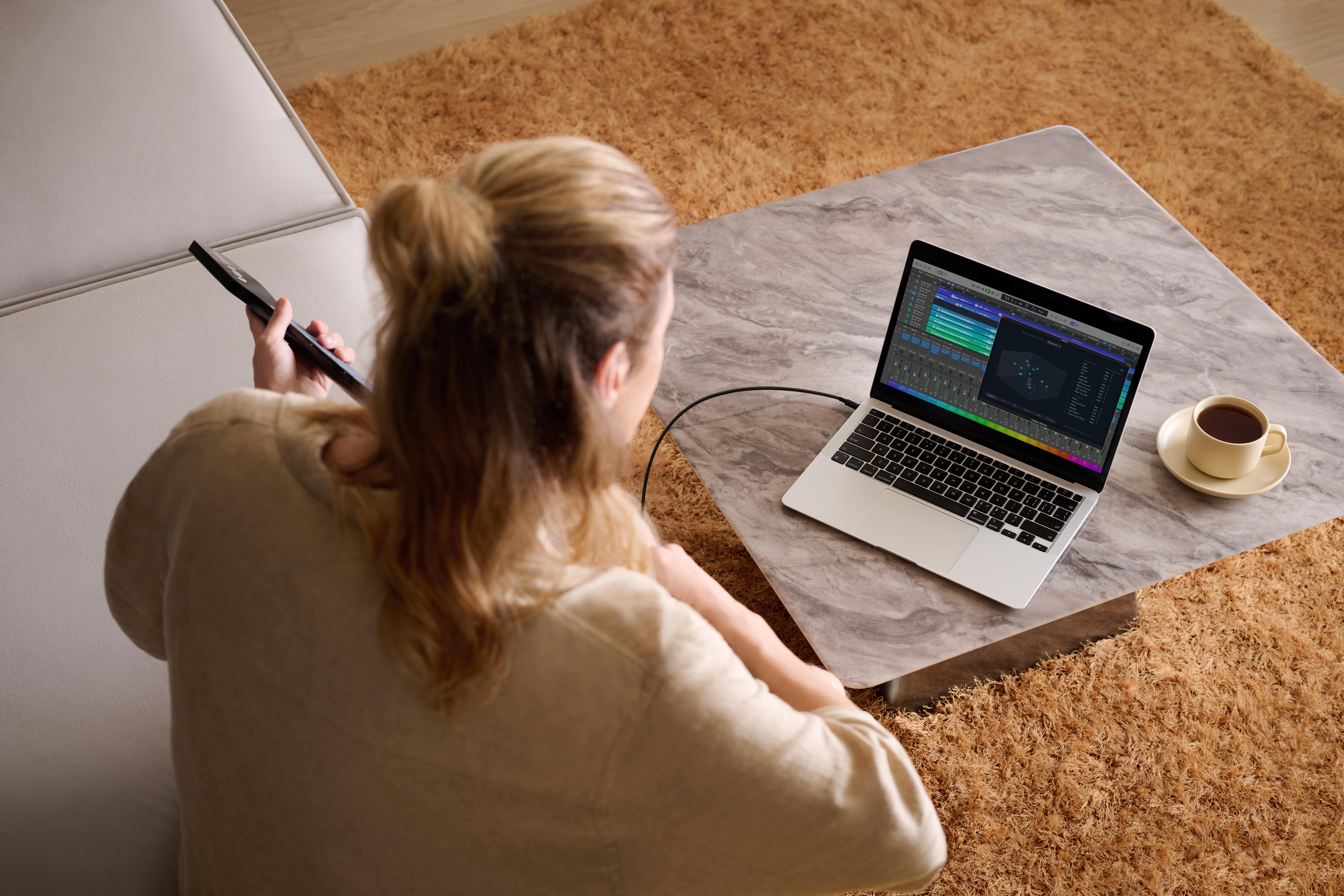Een vrouw met blond haar houdt een afstandsbediening vast. Een laptop staat op een tafel met een kopje koffie. De achtergrond is een shaggy kleed.