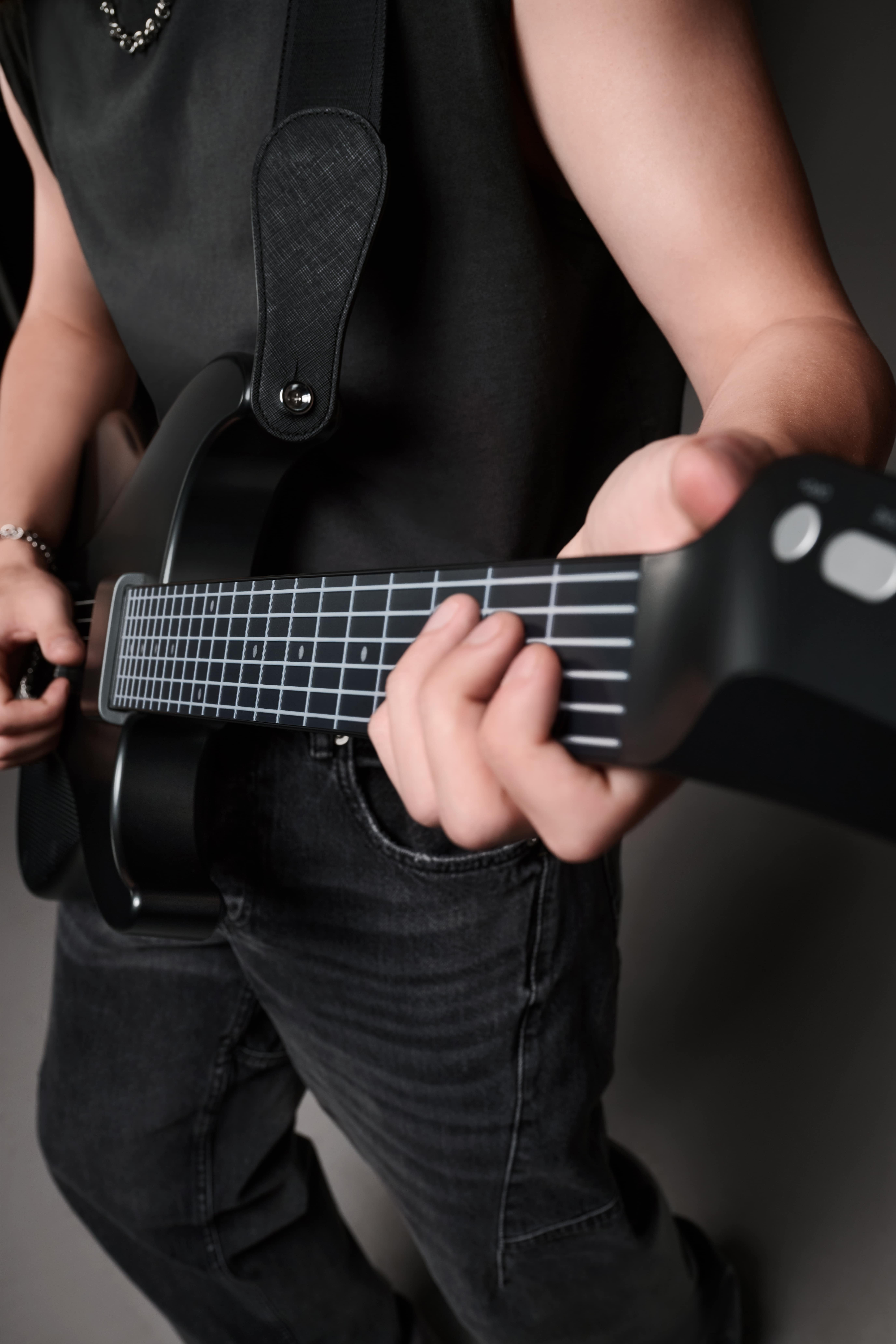 Een persoon speelt op een zwarte gitaar. Ze dragen een zwart shirt en jeans. De achtergrond is neutraal grijs.