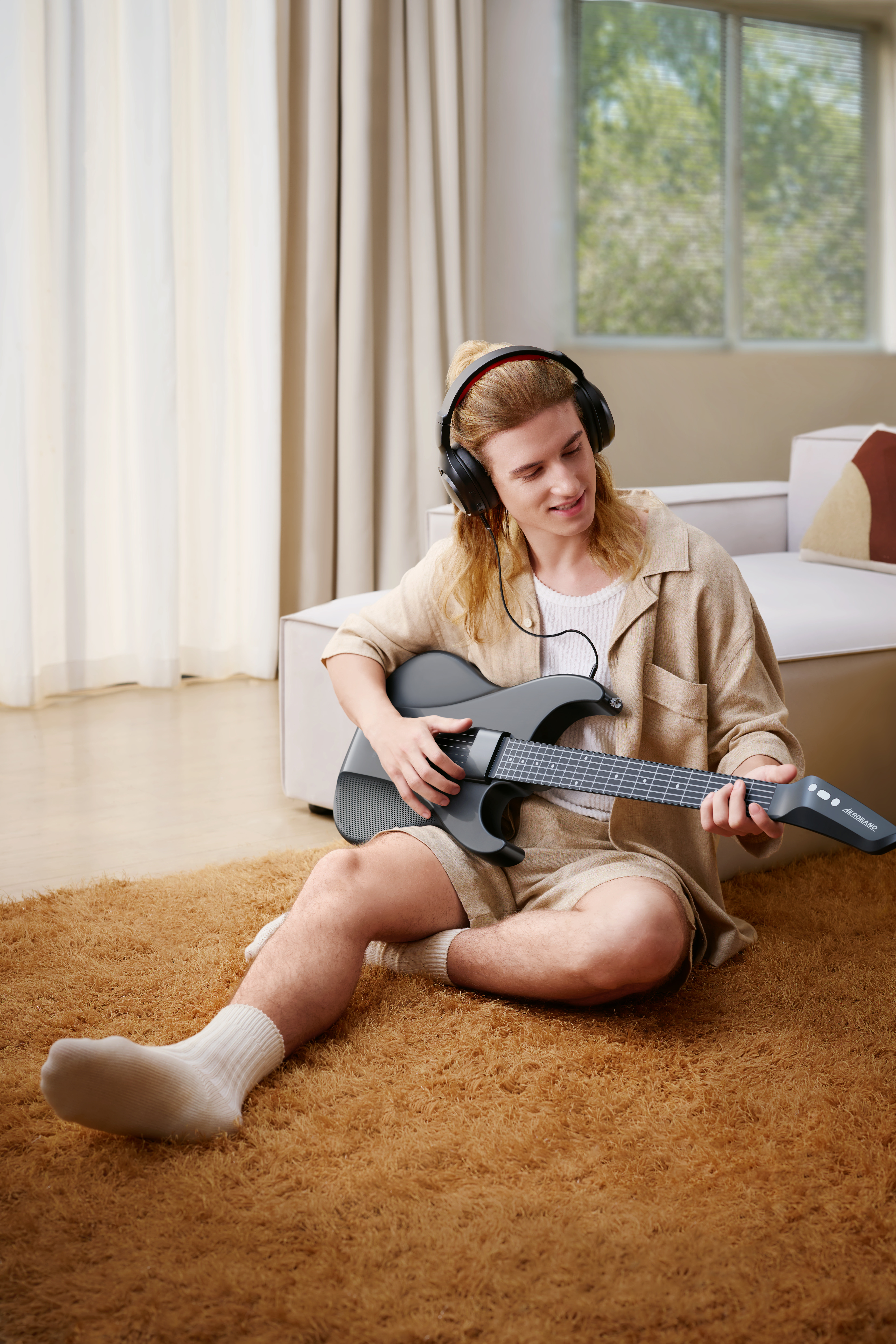 Een man met een koptelefoon speelt gitaar op een bruin tapijt. Hij draagt een korte broek en een shirt.