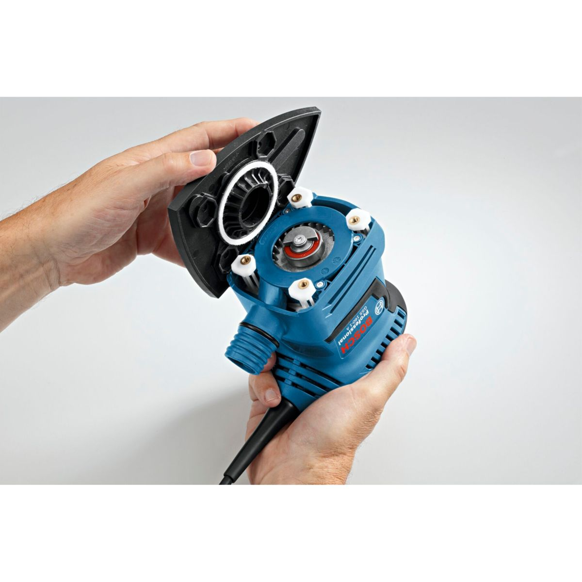 Ręce trzymają niebiesko-czarną szlifierkę, otwierając górną pokrywę. Szlifierka ma białe i czerwone detale. Tło jest białe.