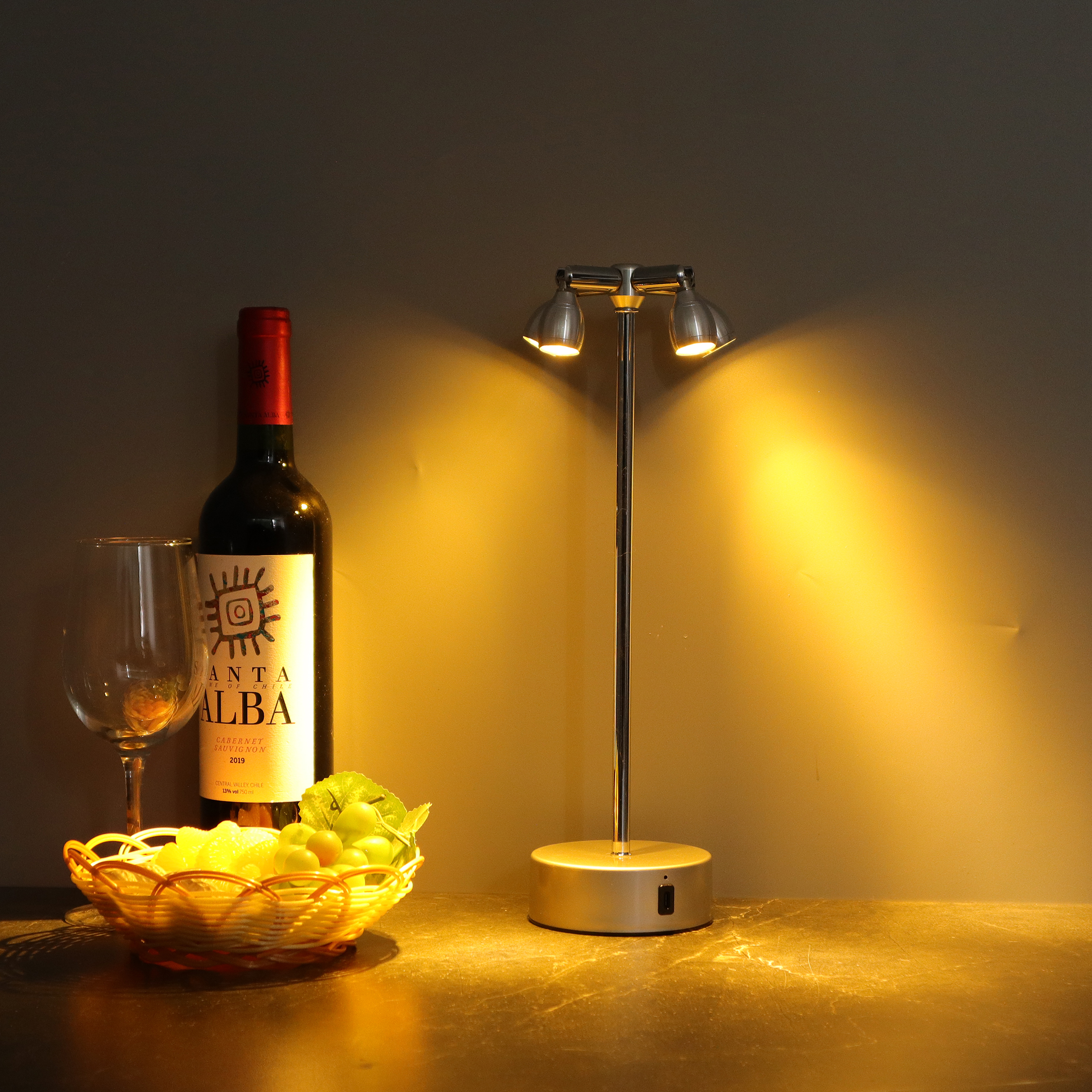 Eine Weinflasche, ein Glas, ein Obstkorb und eine Lampe auf einem Tisch, alles von warmem Licht beleuchtet.