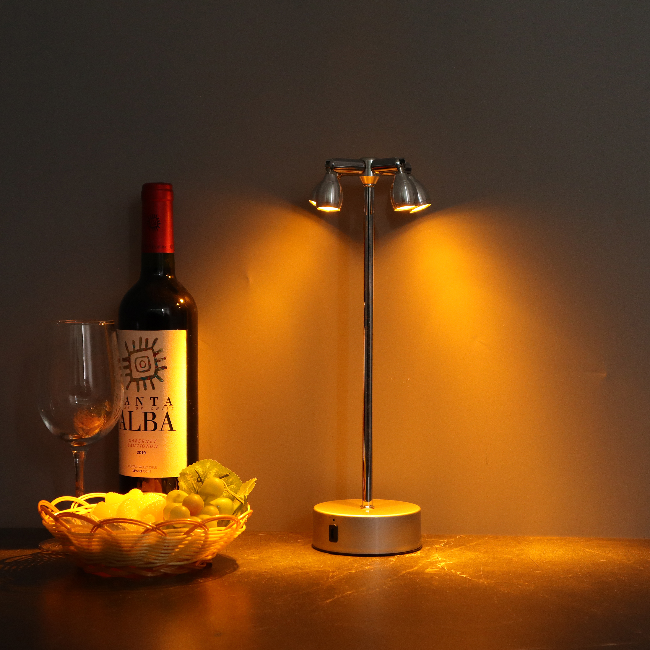 Eine Tischszene mit Weinflasche, Glas, Obstkorb und Lampe. Die Lampe ist beleuchtet.
