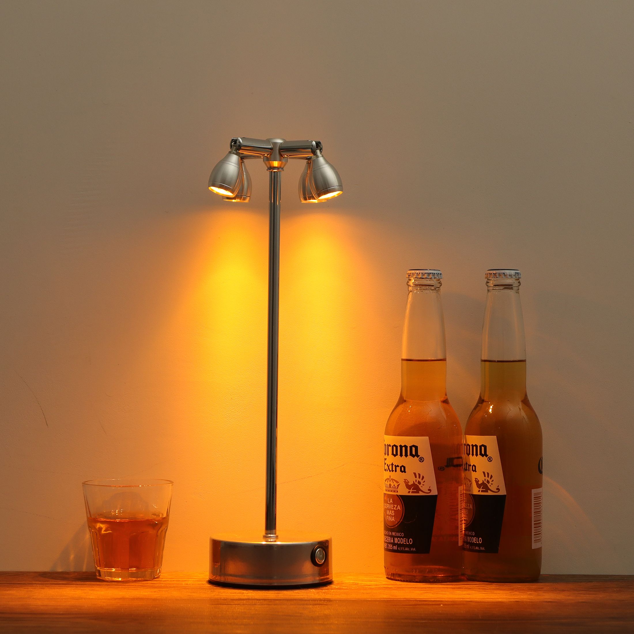 Eine Tischlampe, zwei Corona-Bierflaschen und ein Glas bernsteinfarbener Flüssigkeit stehen auf einer Holzoberfläche.