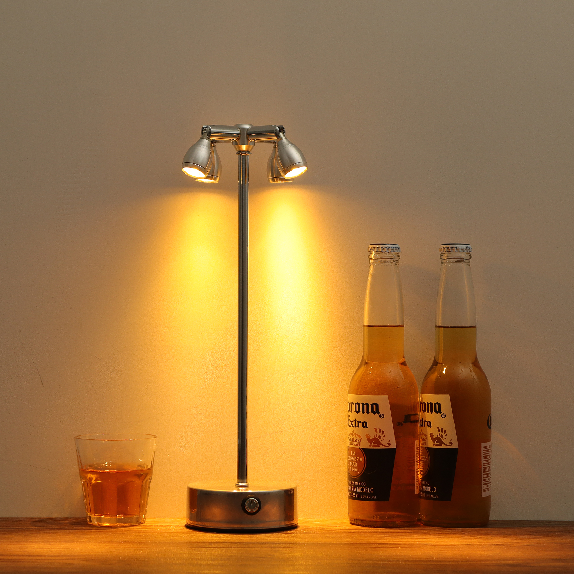 Eine Tischlampe, zwei Bierflaschen und ein Glas bernsteinfarbener Flüssigkeit stehen auf einer Holzoberfläche.