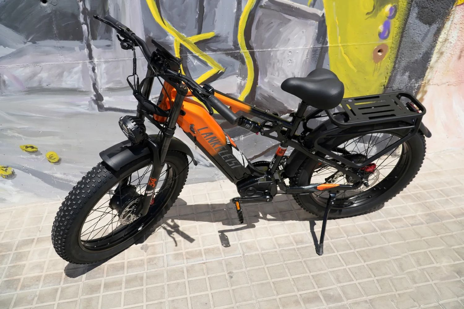 Una bicicleta eléctrica naranja y negra está estacionada sobre una superficie embaldosada. Un muro de graffiti está en el fondo.