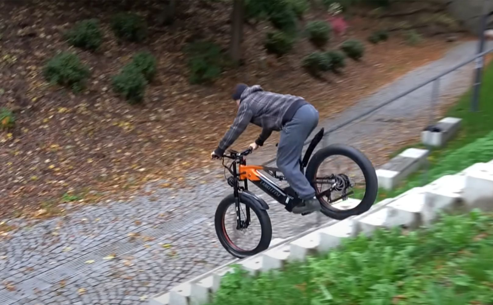 Una persona monta en bicicleta de llantas anchas negra y naranja por unas escaleras. La escena es al aire libre con follaje y un camino de piedra.