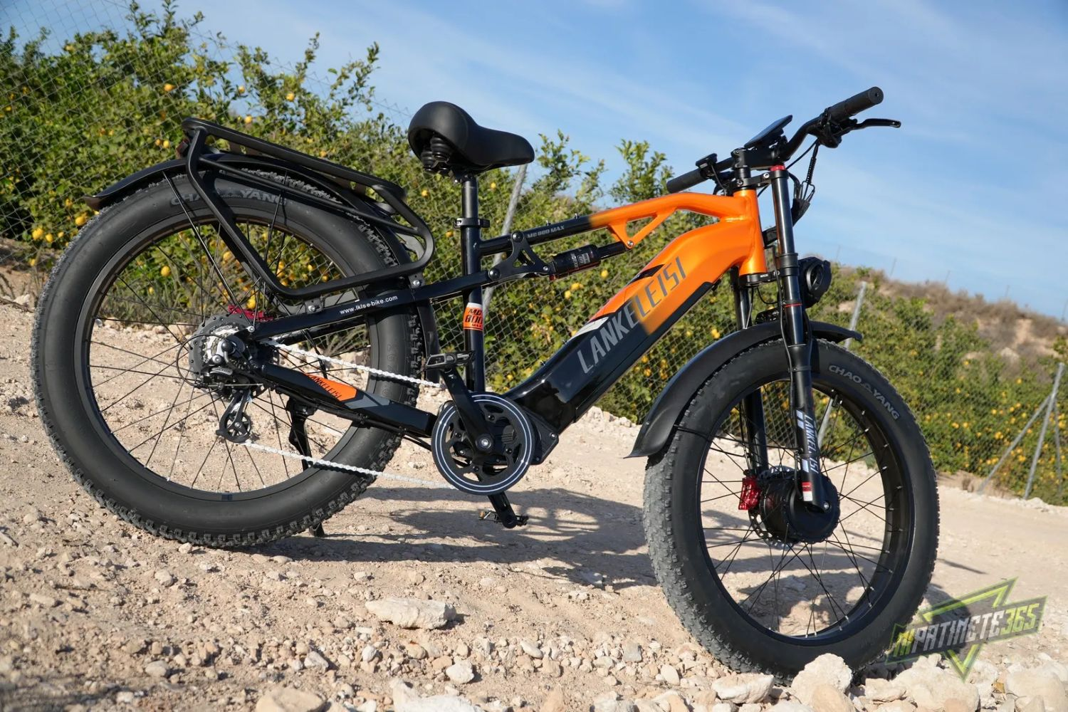 Una bicicleta eléctrica naranja y negra en un camino de tierra con follaje verde y cielo azul de fondo.