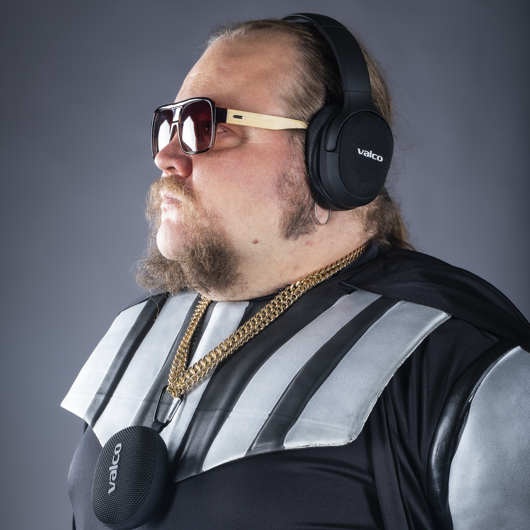 Ein Mann mit Sonnenbrille, Kopfhörern und einer Goldkette. Er trägt auch ein schwarzes Outfit mit silbernen Streifen und hält einen Lautsprecher.