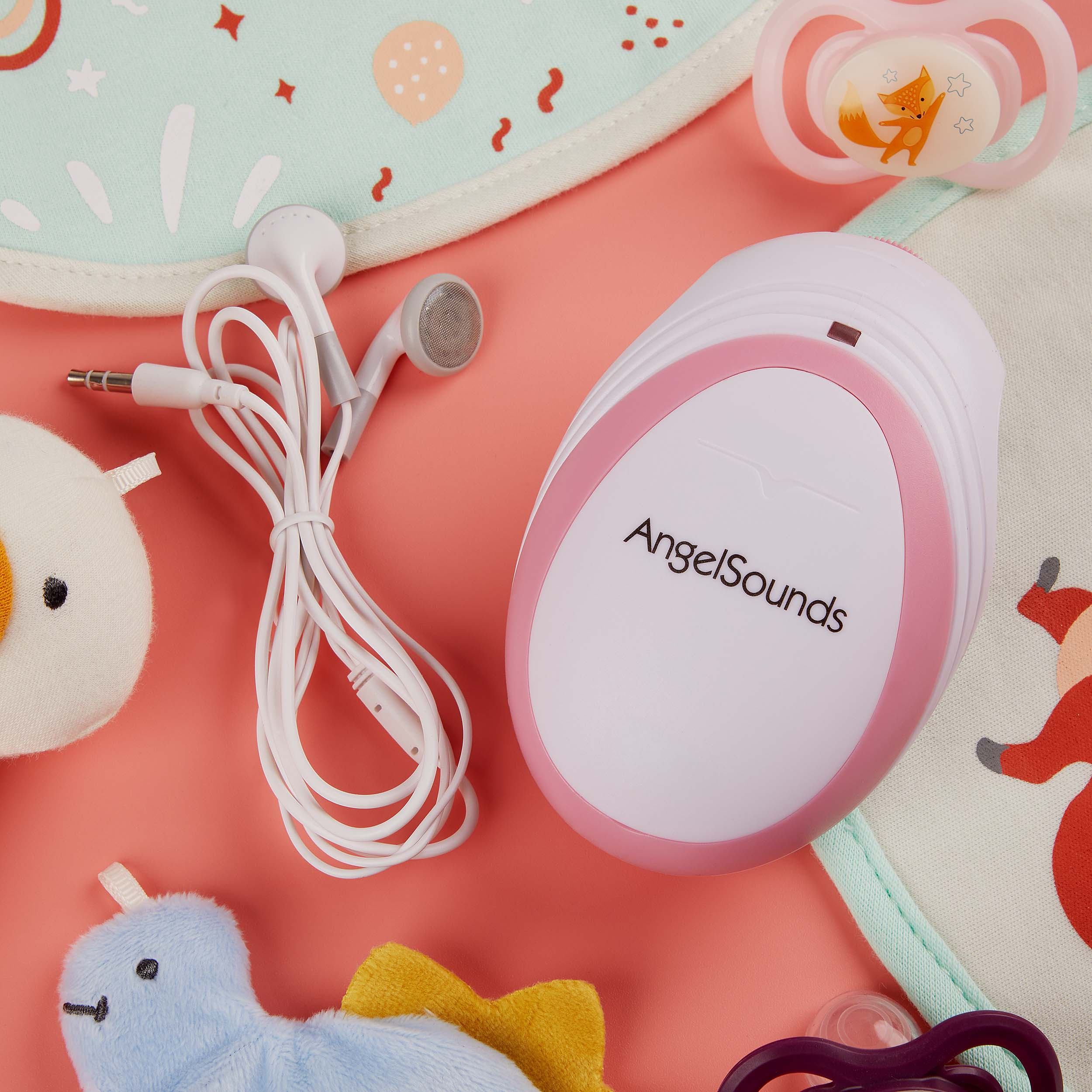 Ein Babyphone, Schnuller, ein Spielzeug und Lätzchen liegen auf einer rosa Oberfläche. Auch Kopfhörer sind vorhanden.