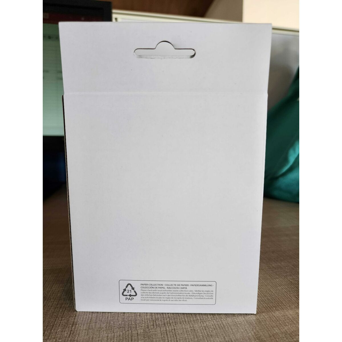 Eine weiße Box mit einem Griffloch. Text unten zeigt Papier- und Recyclinganweisungen. Der Hintergrund ist unscharf.