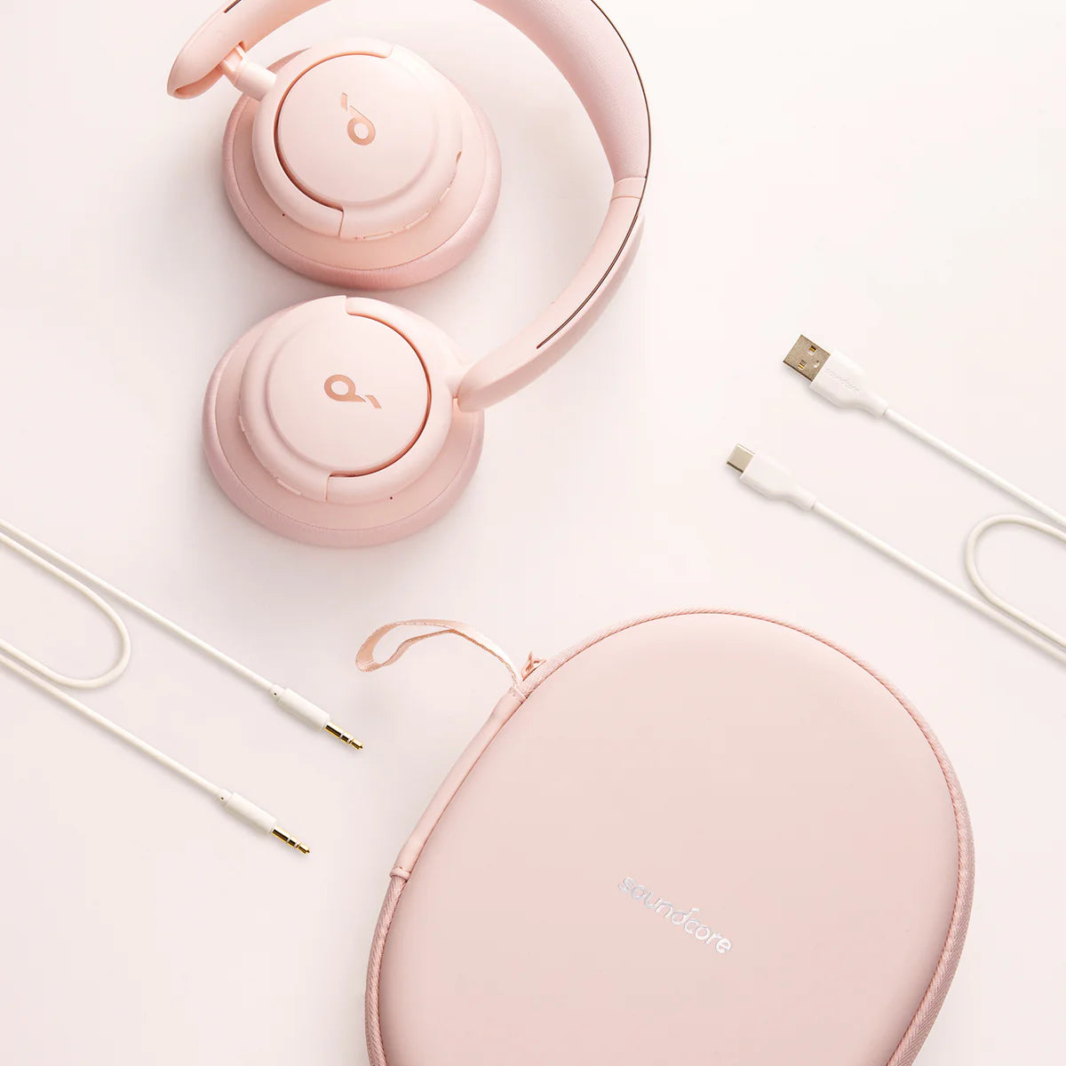Rosa Soundcore-Kopfhörer, Etui und Kabel auf weißem Hintergrund. Kopfhörer sind offen, das Etui oval, die Kabel weiß.