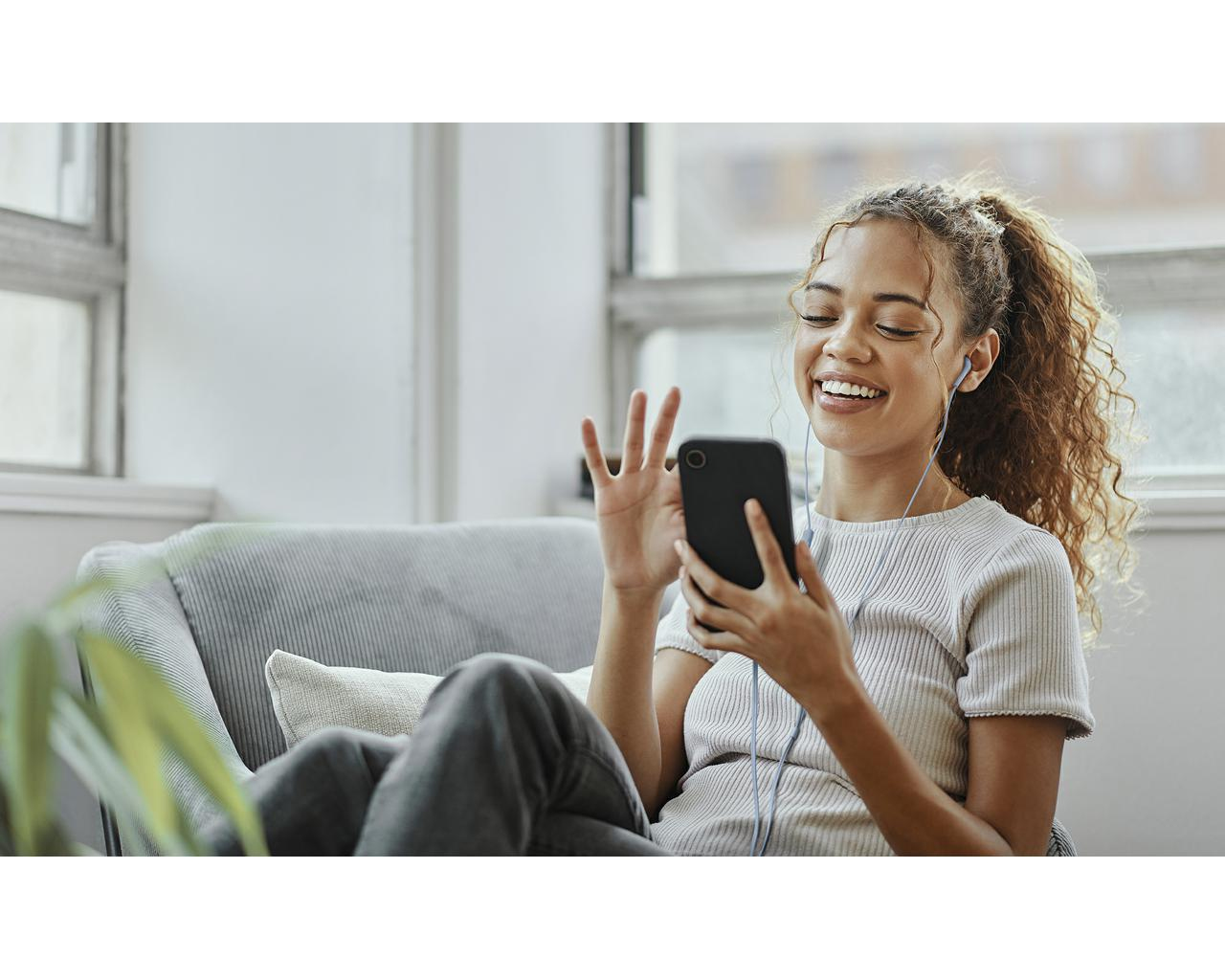 Kobieta z kręconymi włosami siedzi na kanapie, uśmiecha się i trzyma telefon. Ma słuchawki i macha.
