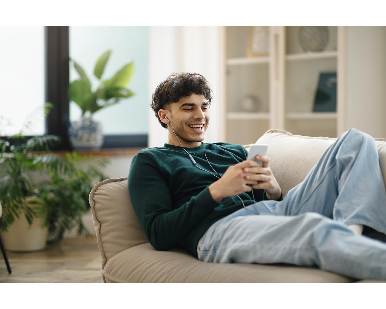 Uśmiechnięty mężczyzna leży na kanapie, używając smartfona. Ma na sobie zielony sweter i dżinsy, ze słuchawkami. Rośliny i regał w tle.