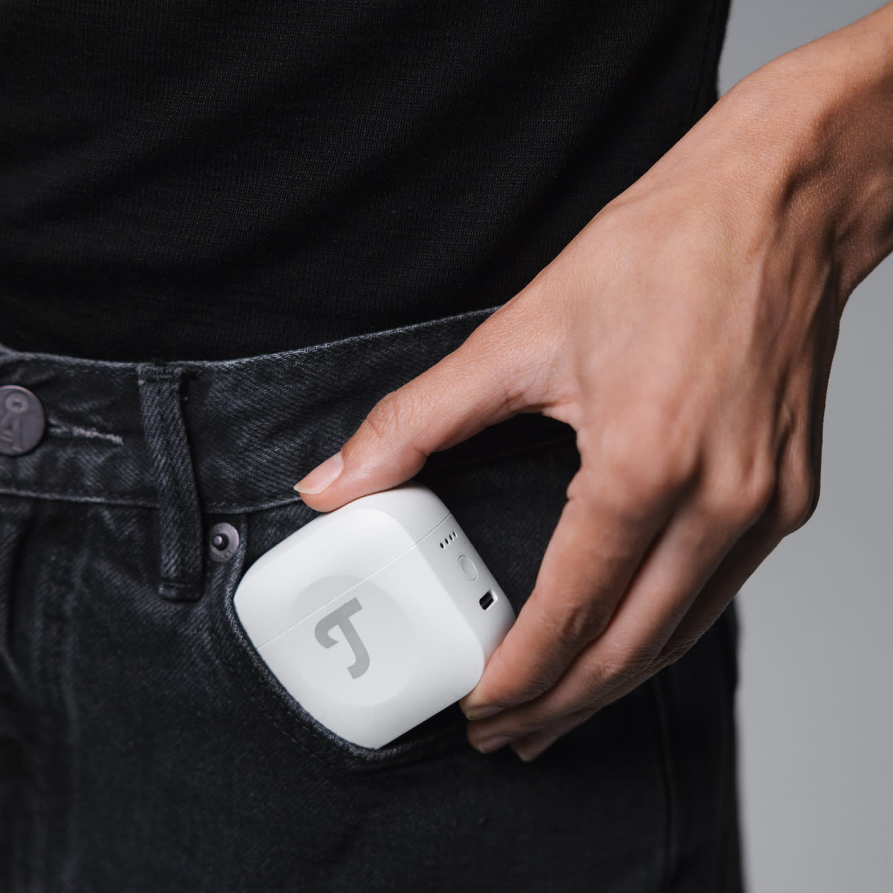 Dłoń trzyma białe kwadratowe urządzenie z logo 'J' w czarnej kieszeni dżinsów, na szarym tle.