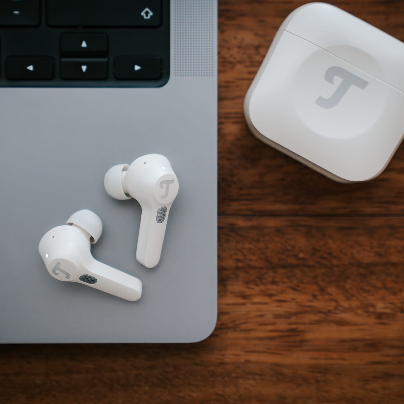 Białe bezprzewodowe słuchawki i etui ładujące na laptopie i drewnianej powierzchni. Słuchawki mają literę 'T'.