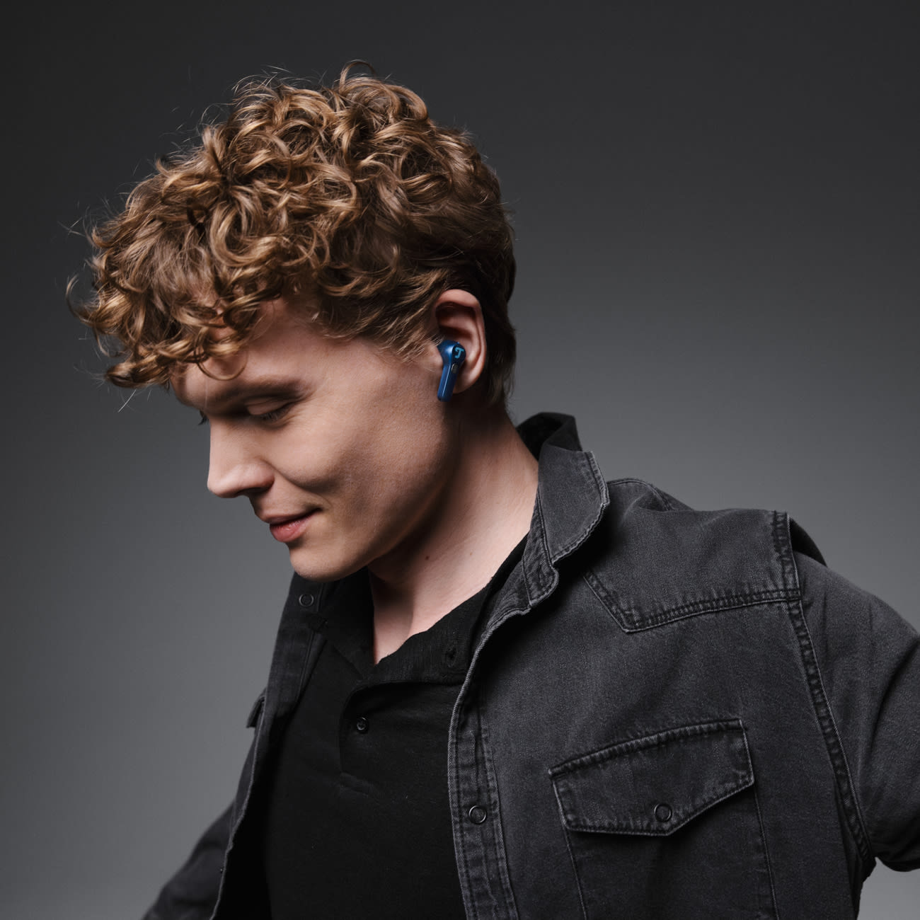 Mężczyzna z kręconymi włosami w niebieskich słuchawkach, czarnej koszuli i dżinsowej kurtce, patrzący w dół. Szare tło.
