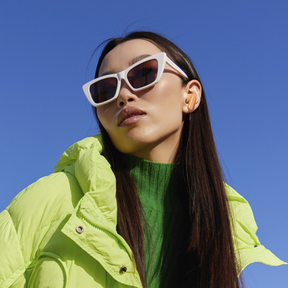 Kobieta w okularach przeciwsłonecznych, zielonym swetrze i neonowej kurtce. Ma wkładki douszne. Tło nieba.