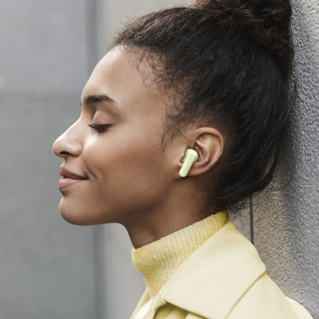 Kobieta z zamkniętymi oczami w żółtym golfie i kurtce, ma słuchawkę. Oparta o szarą ścianę.