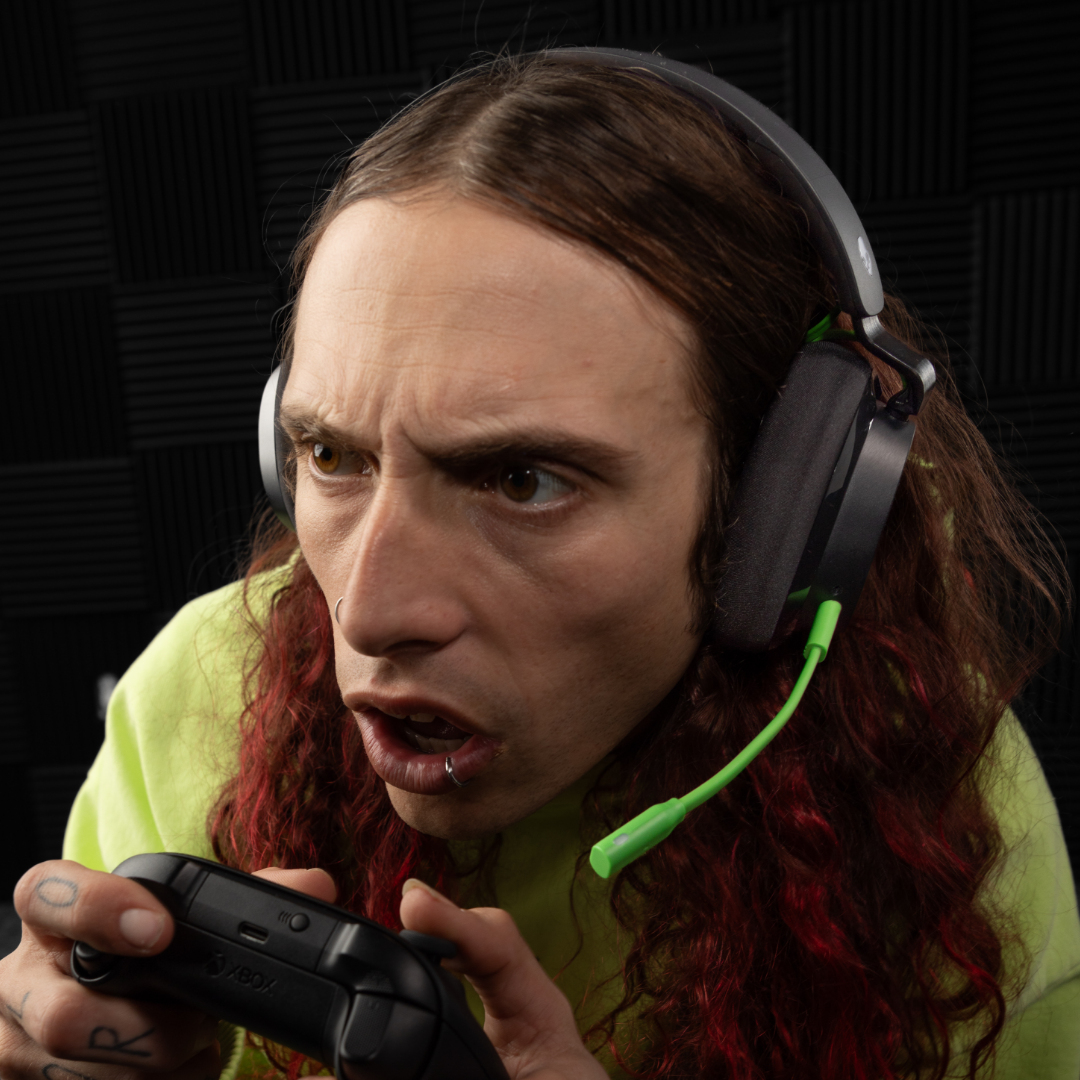Una persona con i capelli lunghi che indossa delle cuffie e tiene in mano un controller di gioco, con uno sguardo concentrato. Lo sfondo è una parete insonorizzata.