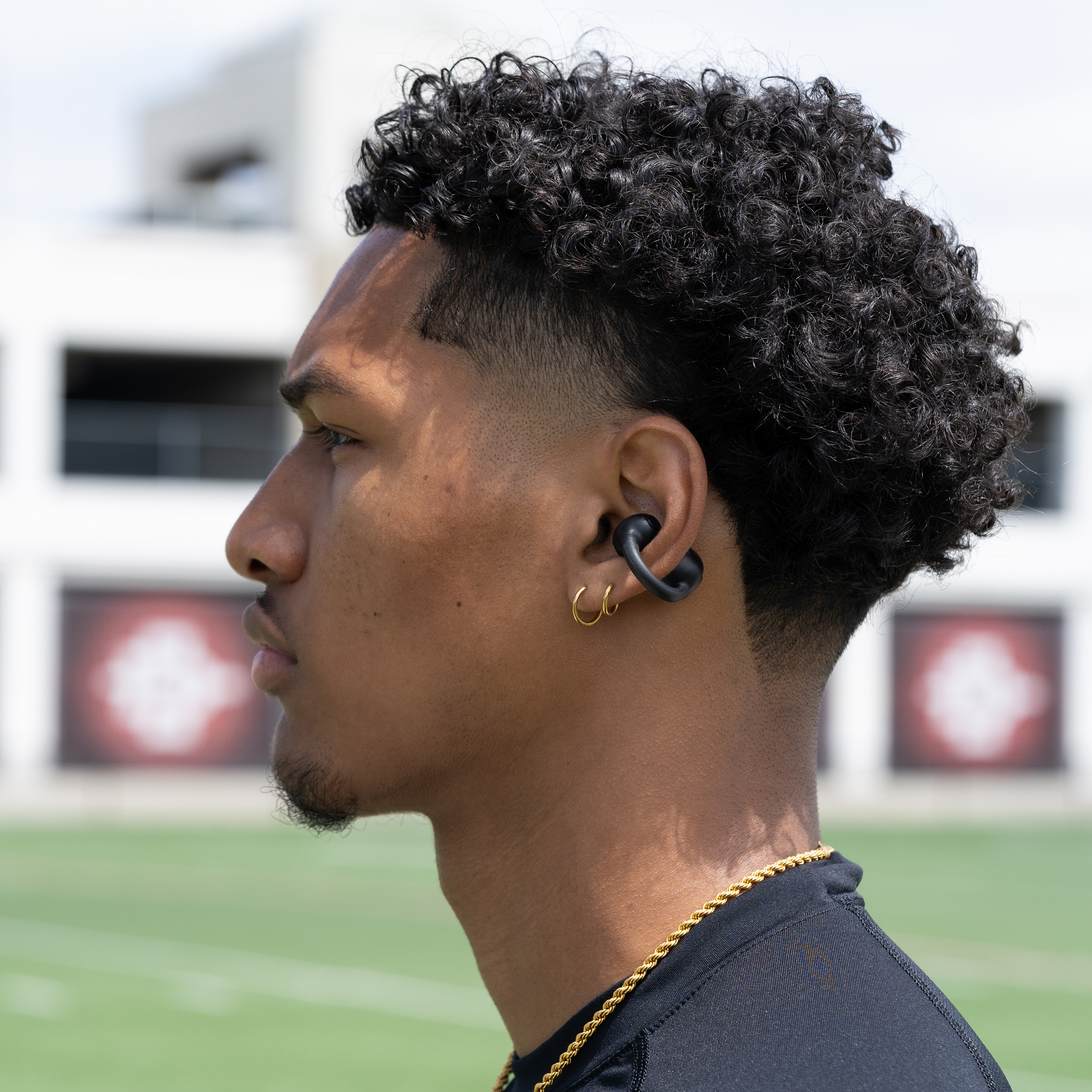 Un homme aux cheveux bouclés porte une chemise noire, une chaîne en or et des boucles d'oreilles. Il a une barbe et un écouteur. Le fond est un champ.