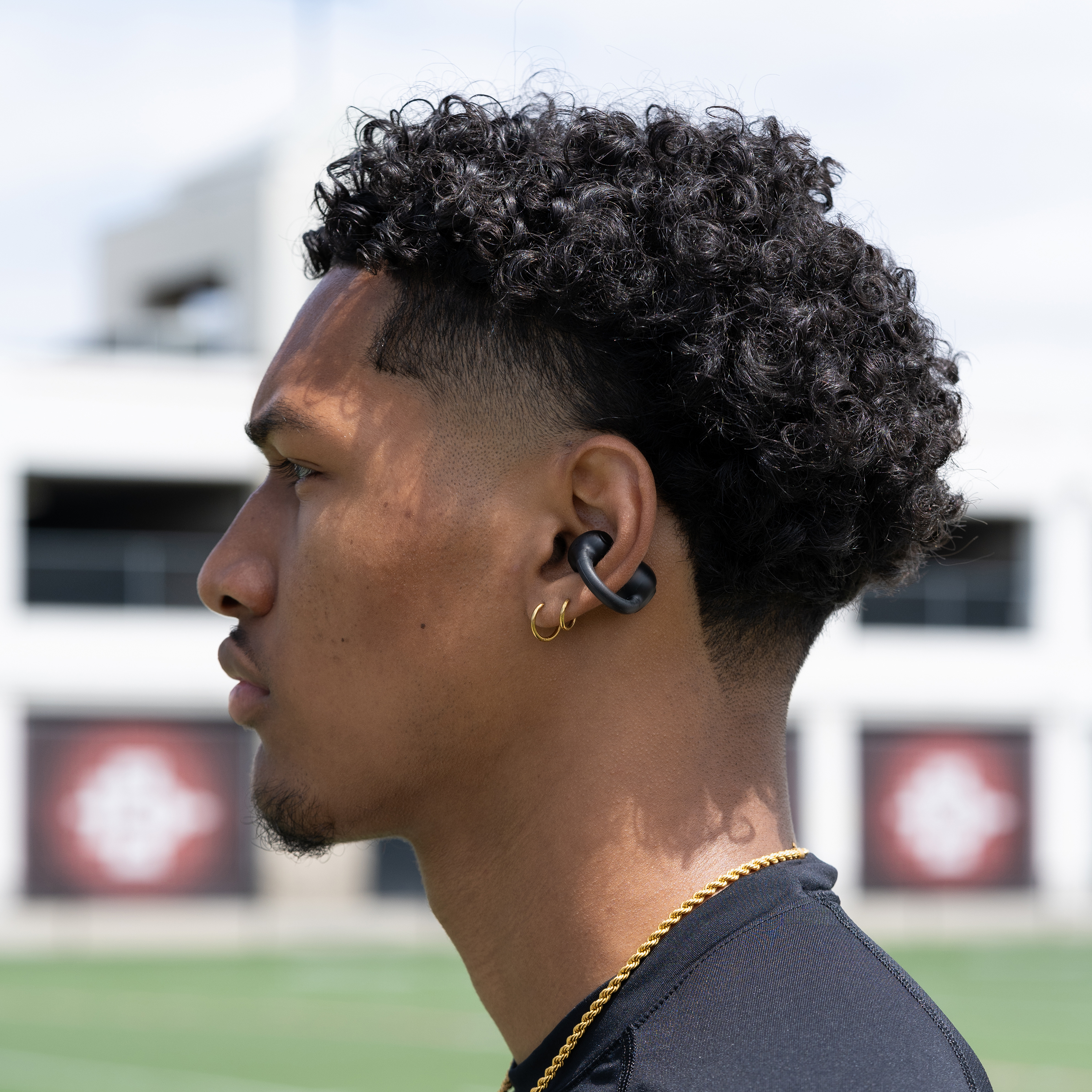 Un homme aux cheveux bouclés porte une chemise noire, une chaîne en or et des boucles d'oreilles. Il a un écouteur noir.
