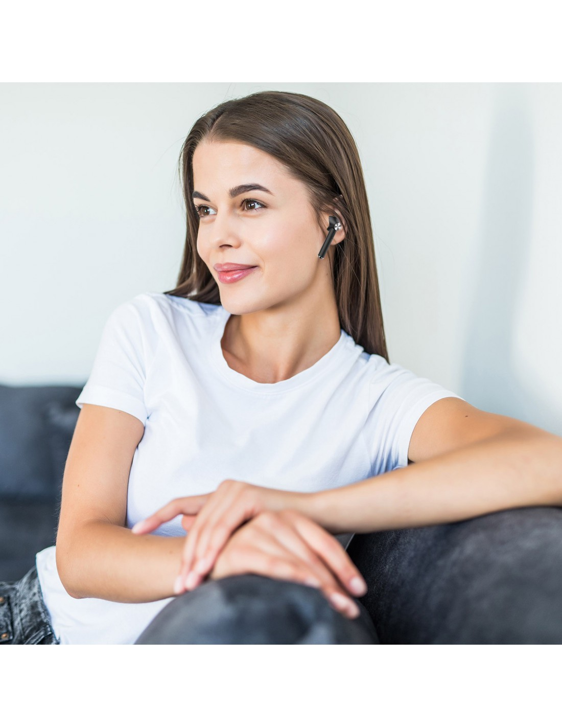 Une femme aux longs cheveux bruns portant un t-shirt blanc, assise sur un canapé gris, portant un écouteur noir. Elle regarde ailleurs.